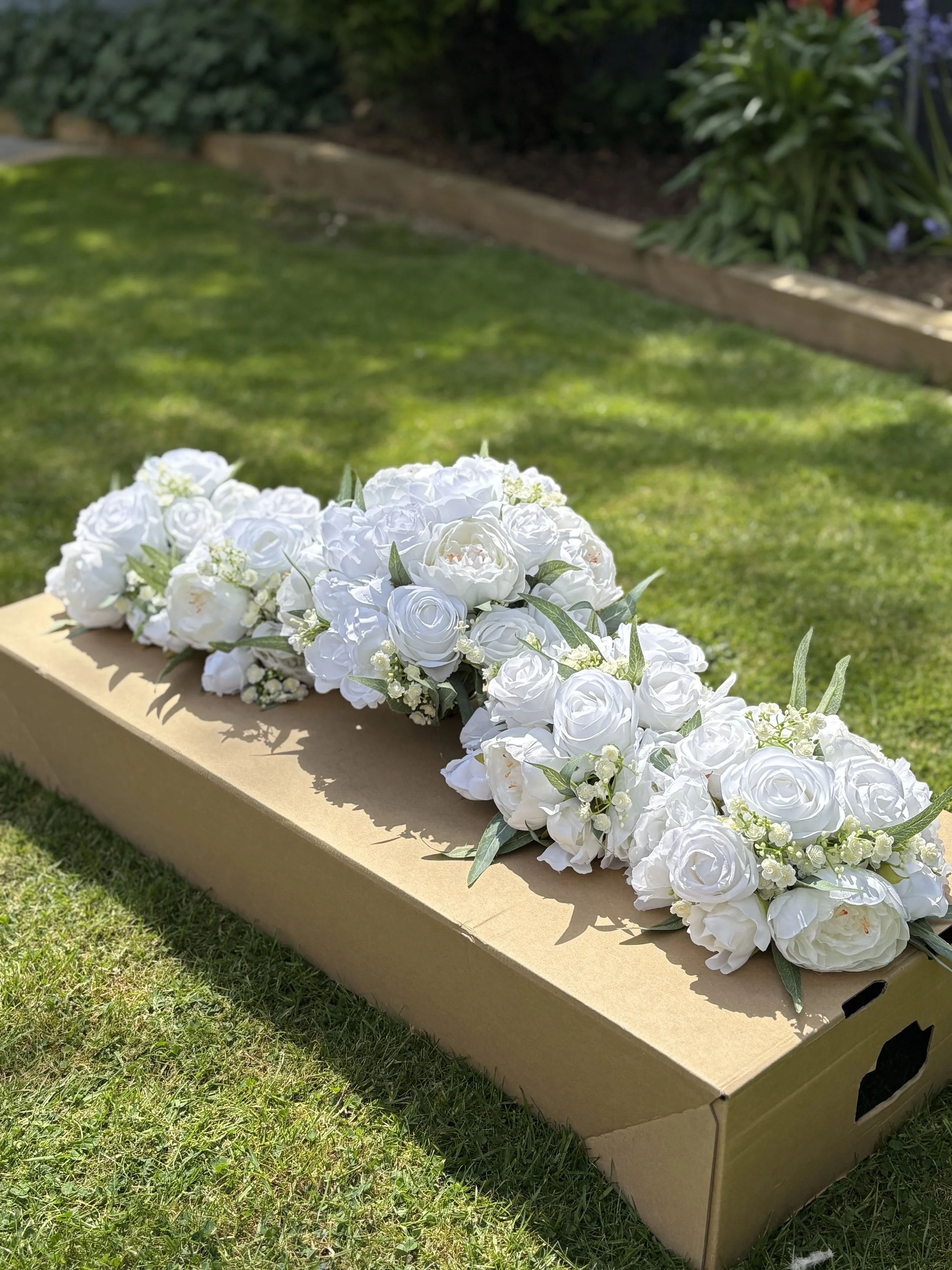 A cardboard box with a white floral arrangement on top placed outdoors on grass, with a garden border and plants in the background.