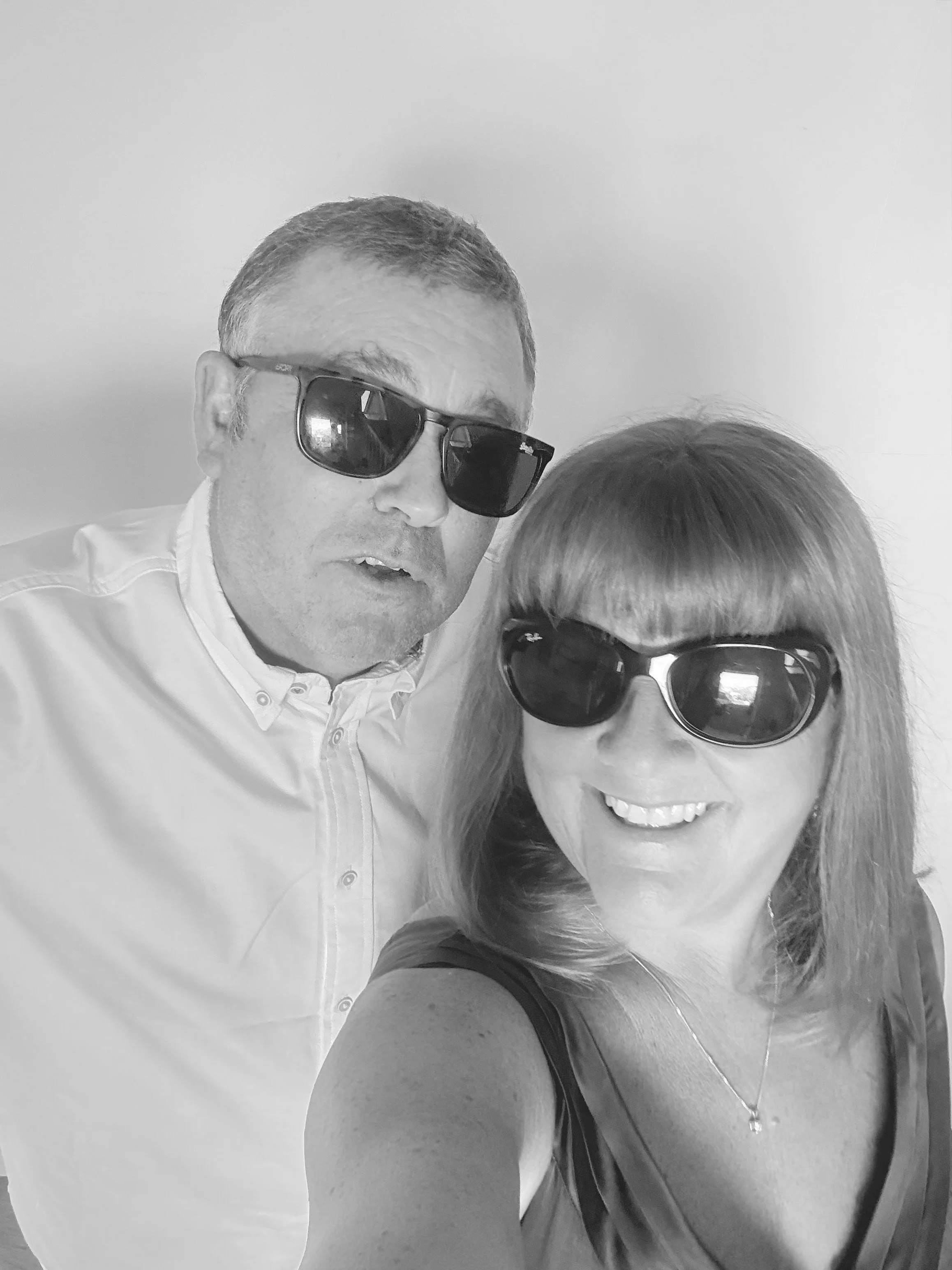 A man and woman taking a selfie together, both wearing sunglasses. The woman is smiling and the man has a neutral expression. Both are dressed casually, with the man in a light-colored button-up shirt and the woman in a sleeveless top.