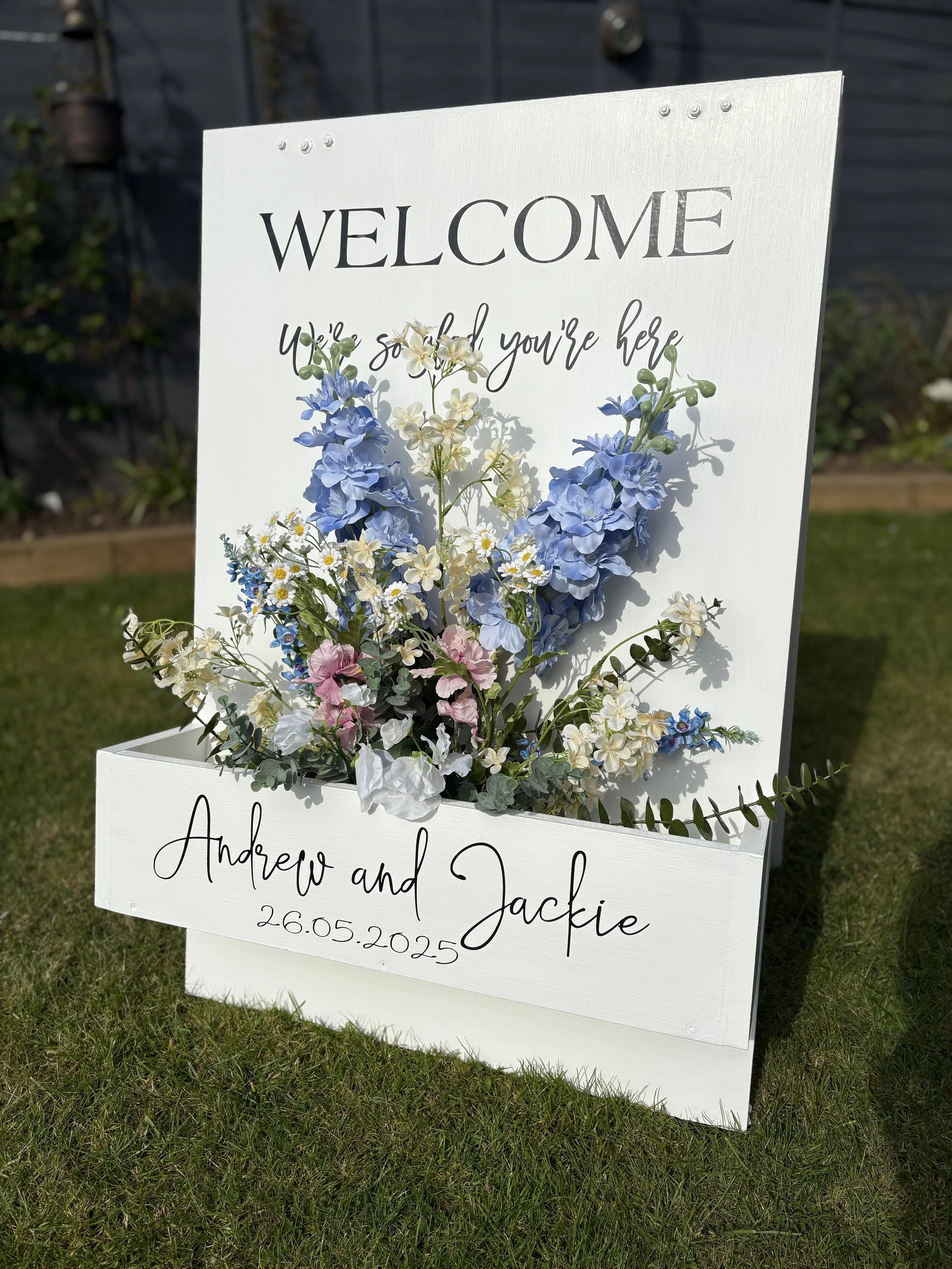 A welcome sign with flowers and personal details at an outdoor event.
