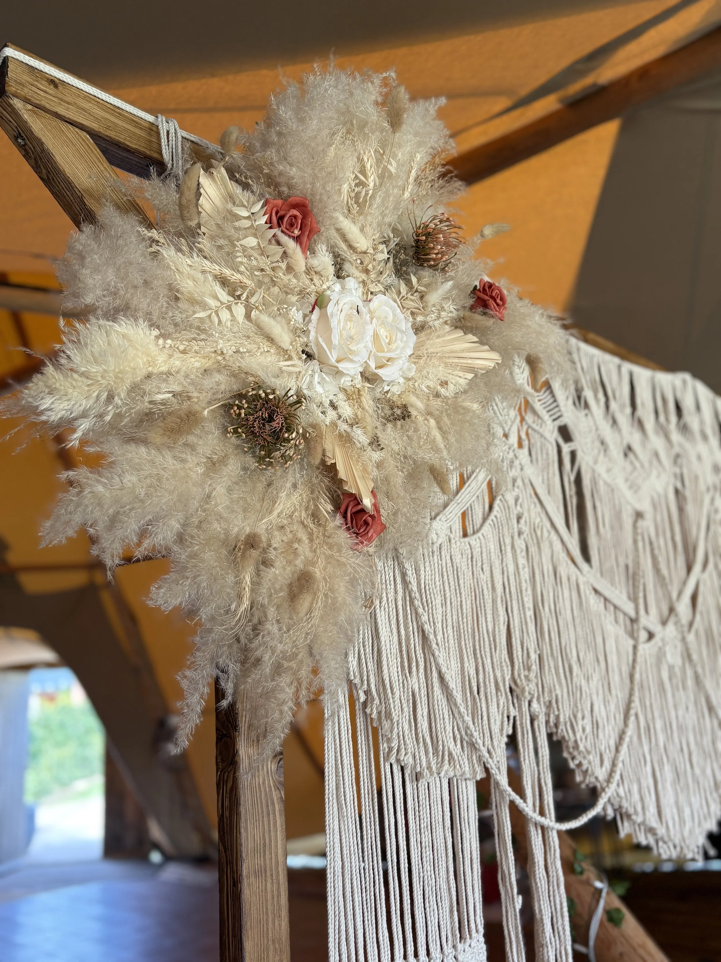 Decorative arrangement of dried flowers and plants with white and pink roses and a macramé wall hanging in the background.