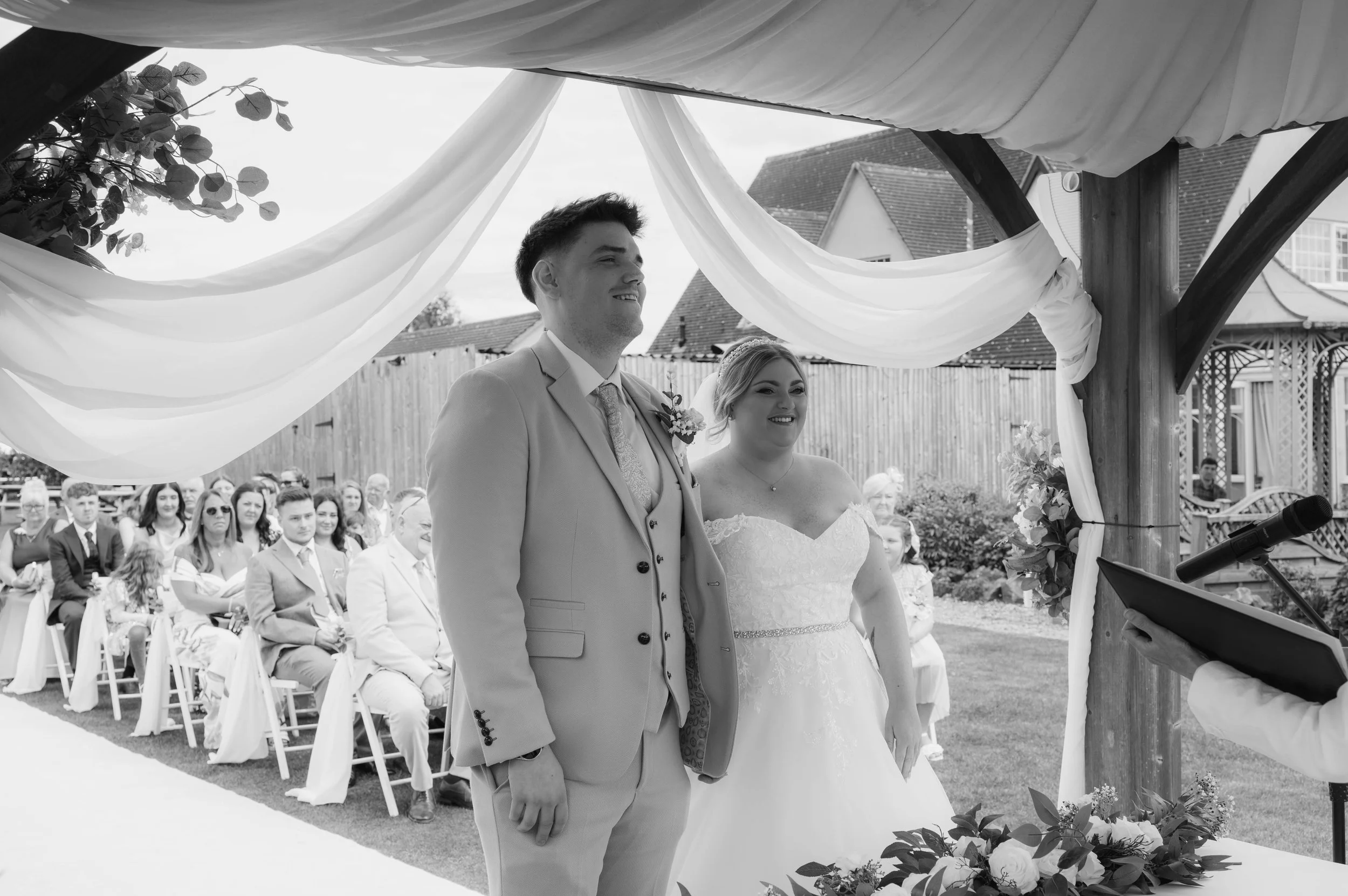 A black and white photo of a wedding ceremony with the bride and groom standing under a decorated wooden arch, with guests seated in the background.