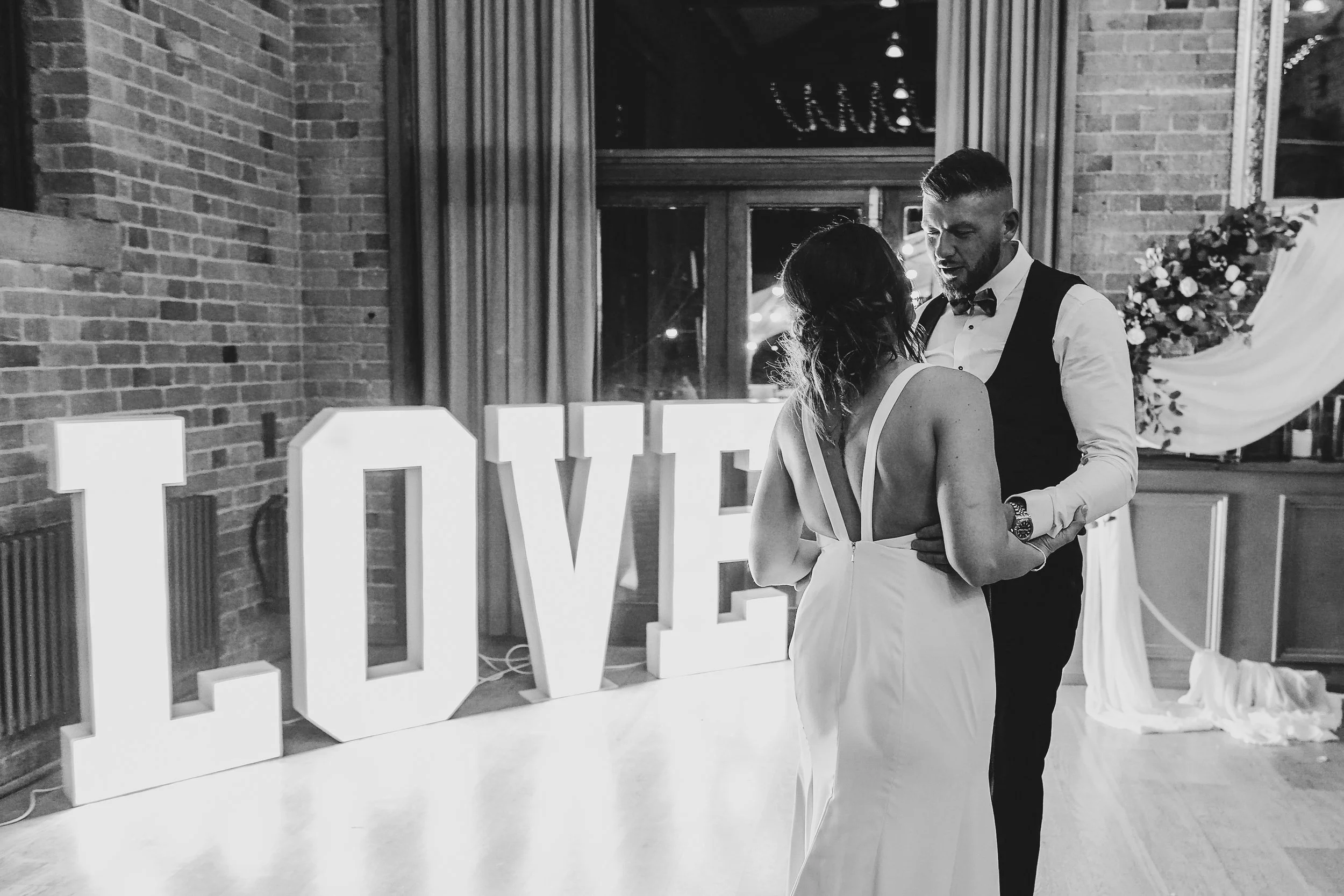A couple dancing together at a wedding reception, with large illuminated letters spelling 'LOVE' in the background, and decorated with flowers and fabric drapery.