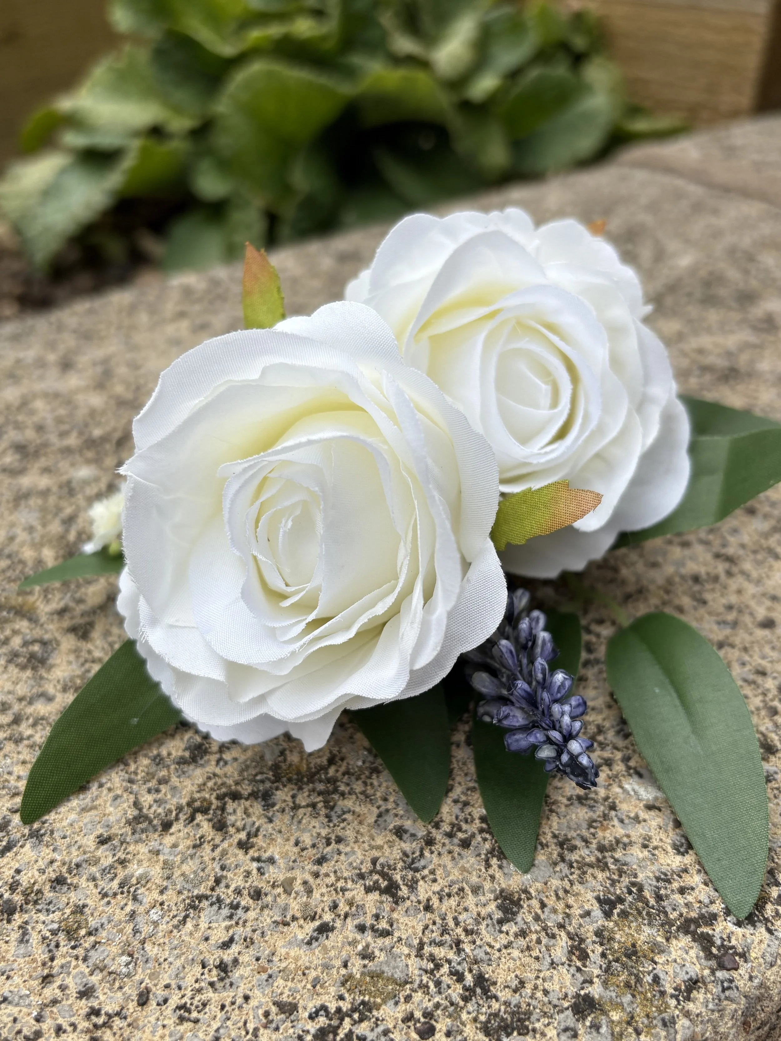 Artificial white roses with green leaves and small purple flowers on a stone surface.