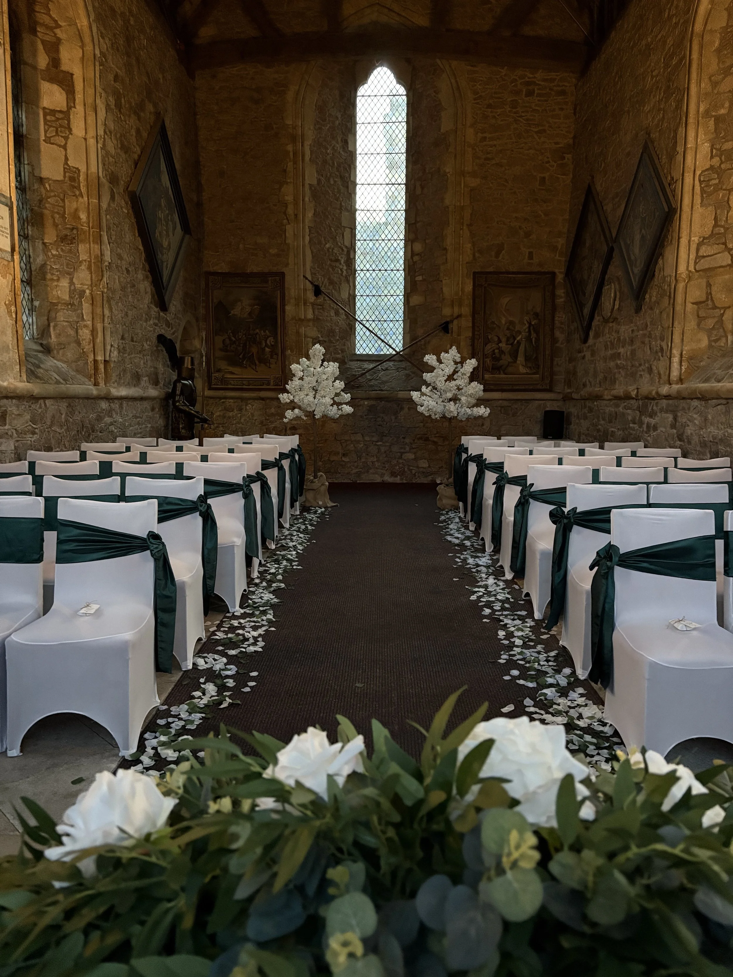Wedding ceremony setup inside a stone church with white chairs, green sashes, flower petals on the aisle, and white floral arrangements at the end of the aisle.