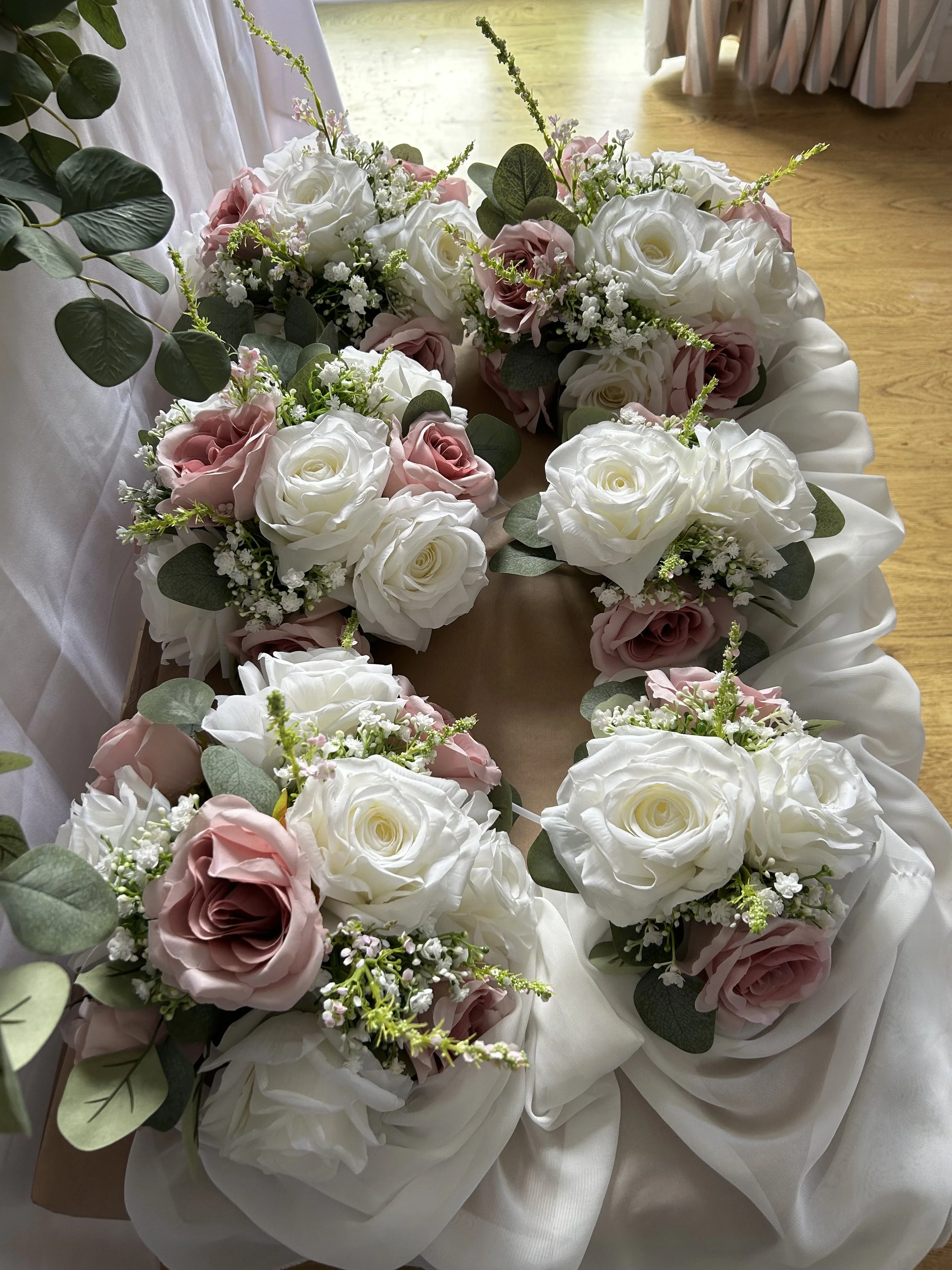 A floral arrangement in the shape of a heart made with white and pink roses, green leaves, and small white filler flowers, placed on a table with white fabric.