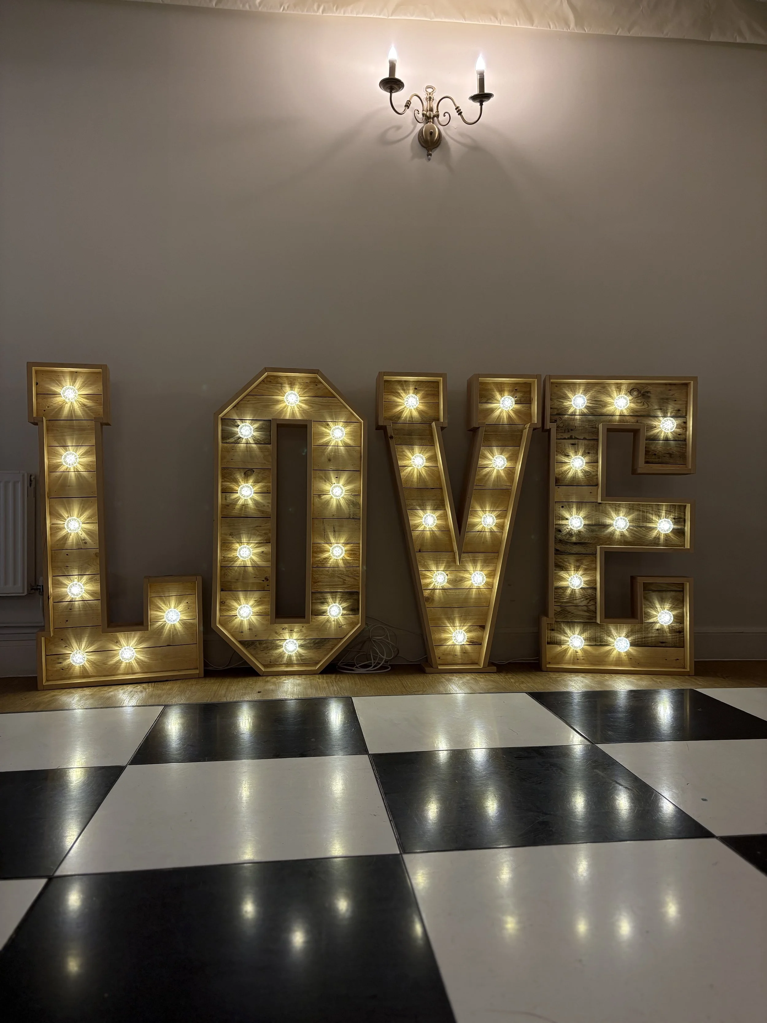 Large illuminated sign spelling out the word 'LOVE' with warm yellow lights, placed against a plain wall on a checkered black and white floor, with a small chandelier hanging from the ceiling above.