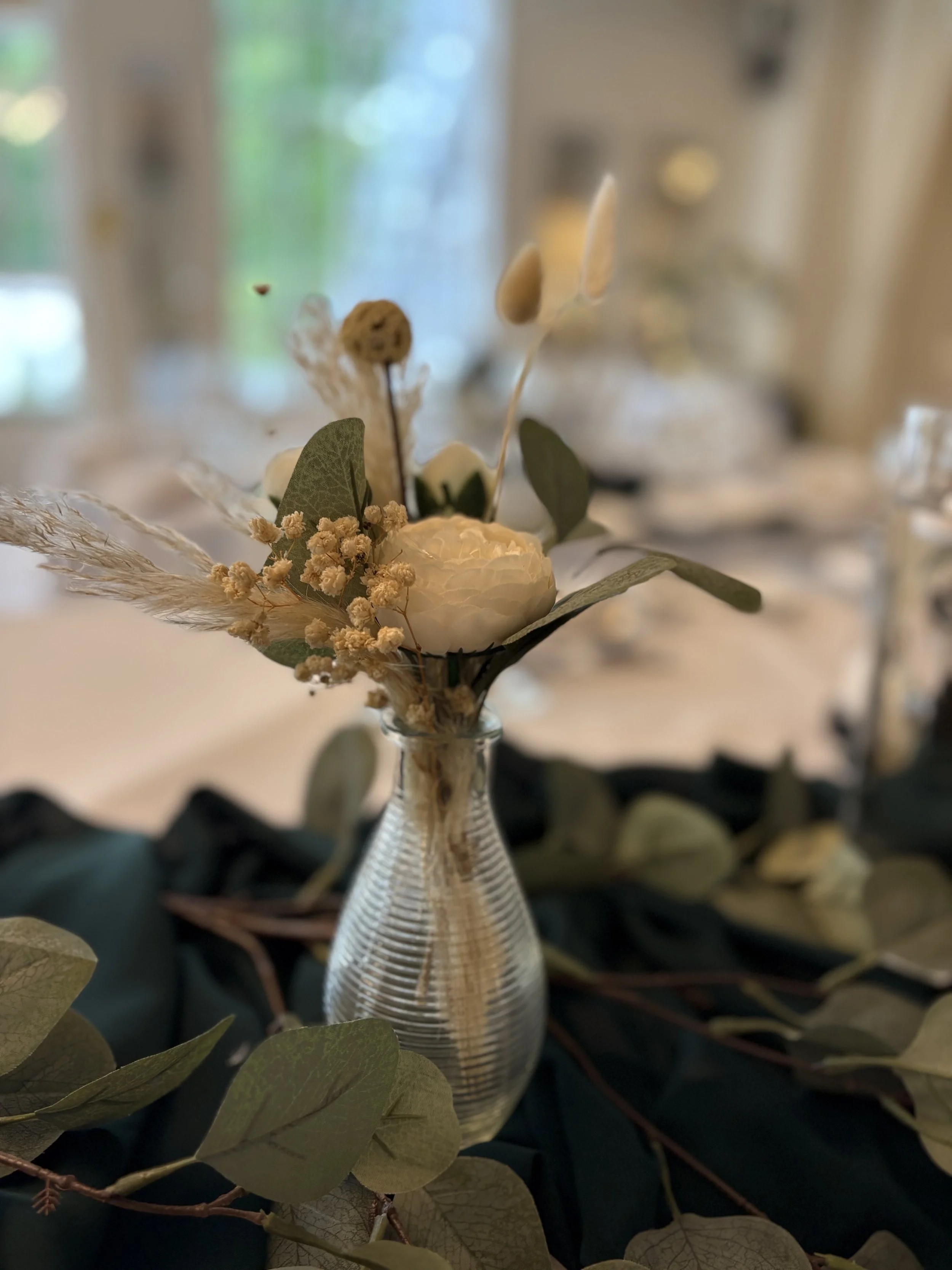 A small clear glass vase holding a decorative arrangement of white and beige dried flowers, with green leaves and natural elements, placed on a dark fabric or foliage surface.