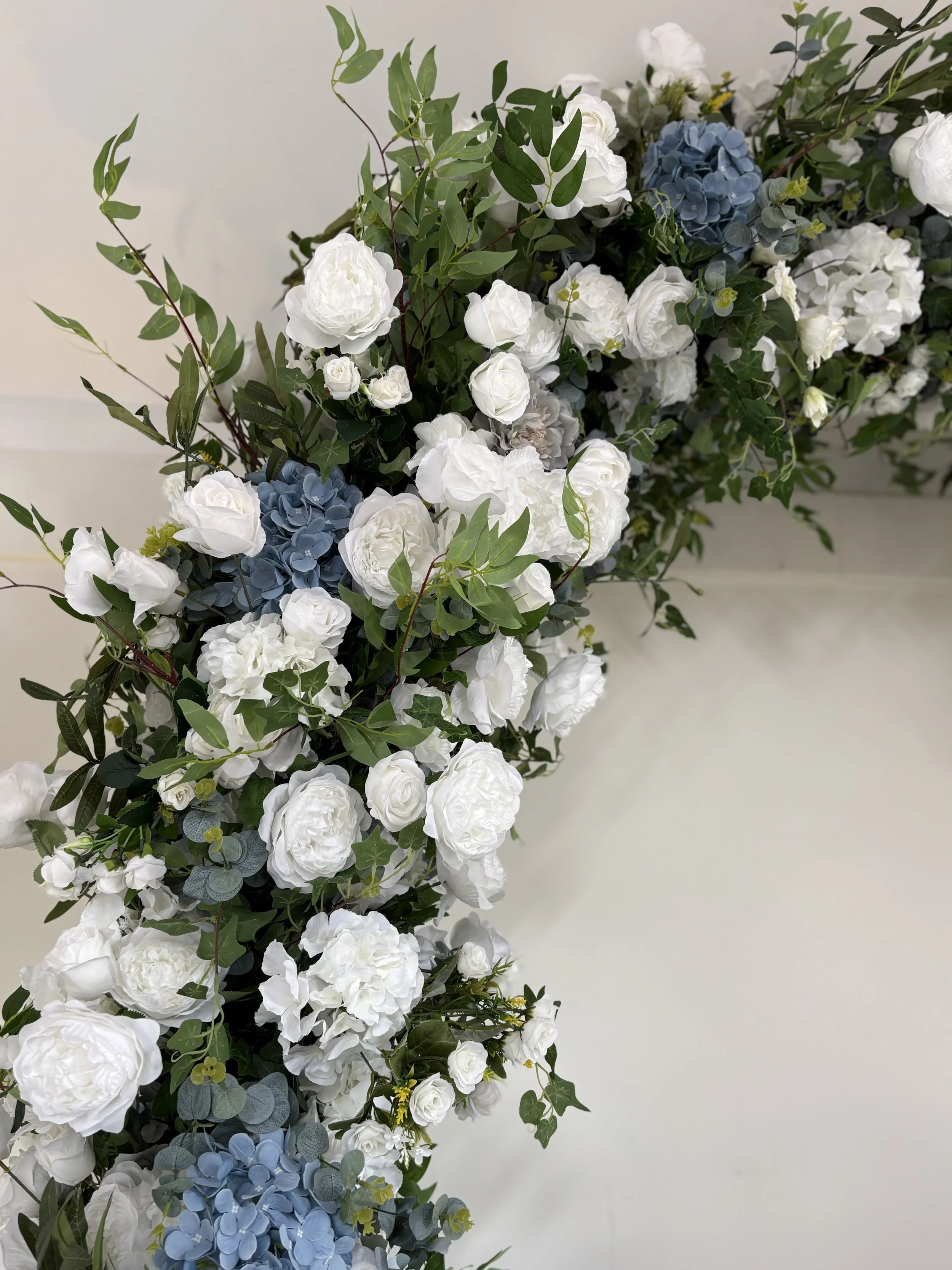 A floral arrangement with white roses, intertwined with green leaves and blue hydrangeas, displayed against a white background.