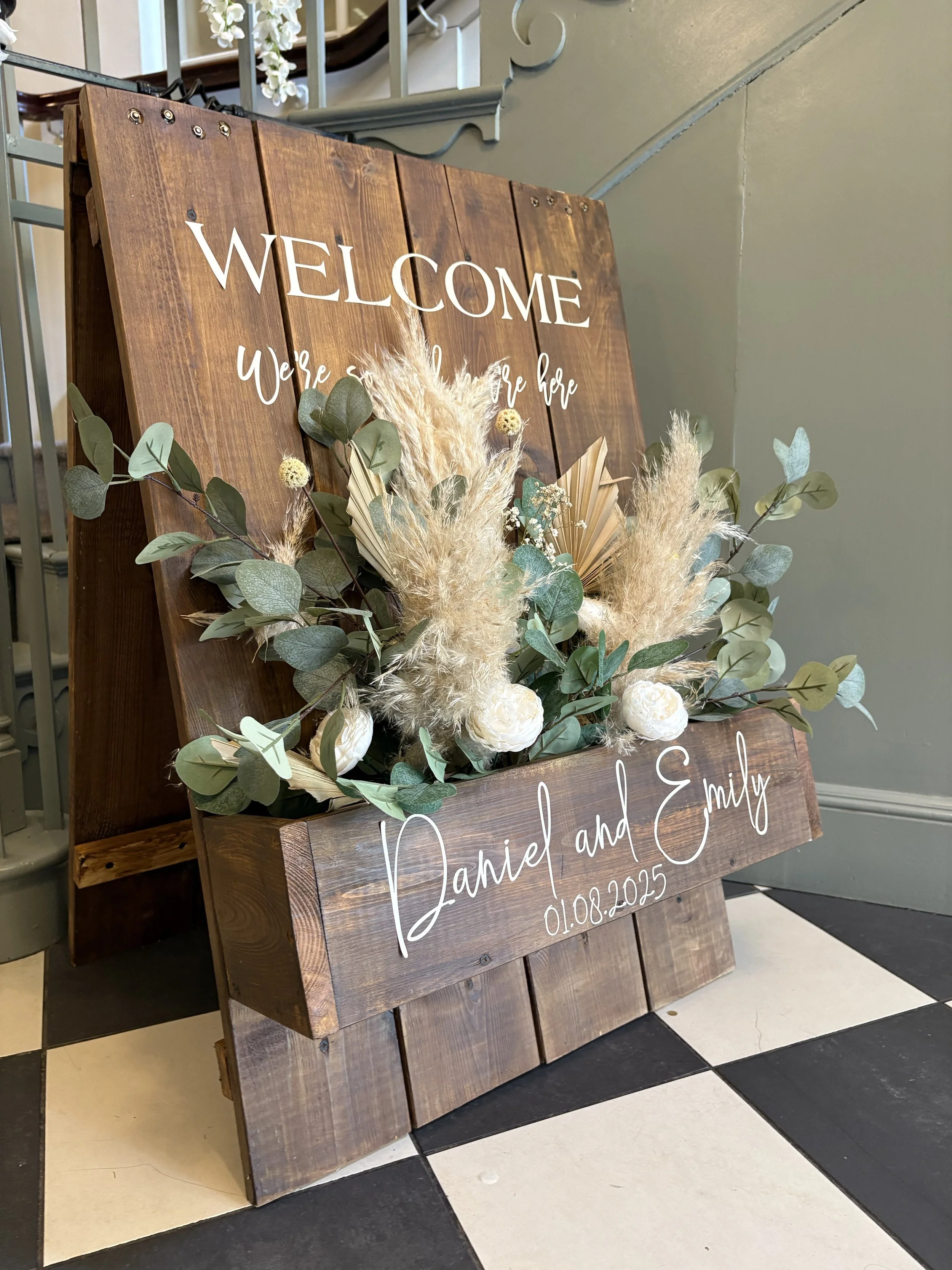 A wooden wedding sign with white lettering welcomes guests and displays the names Daniel and Emily, along with the date 01.08.2025, decorated with greenery, white flowers, and dried pampas grass.