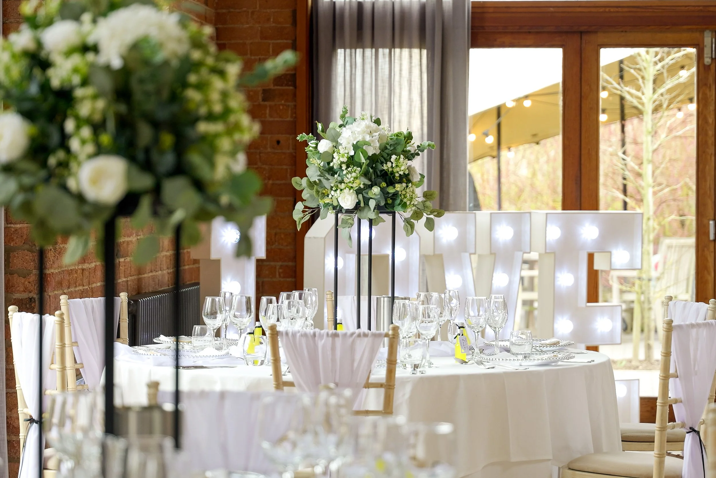 Wedding centrepiece with green and white artificial flower arrangement on a black open lantern  with giant LOVE letters in the background