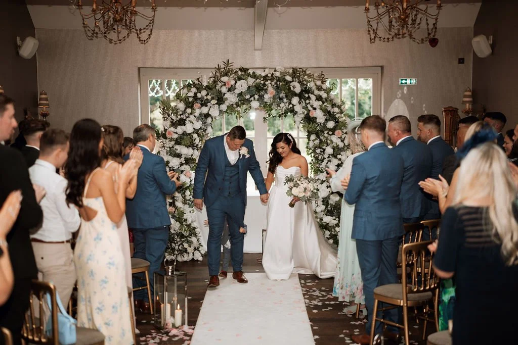 A couple holding hands and walking down the aisle during their wedding ceremony surrounded by friends and family in a decorated indoor venue.