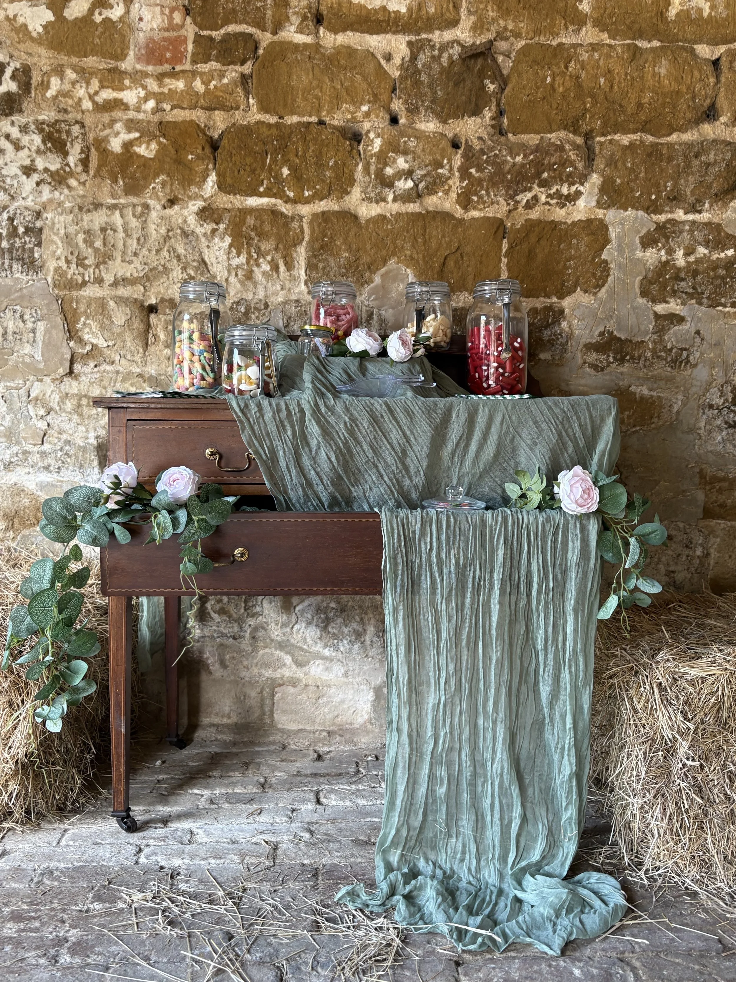 A vintage wooden table decorated with a greenish fabric runner, artificial flowers, and greenery. Behind it, a stone wall with jars filled with colorful candies and treats on top. Hay buffs are placed on either side of the table.