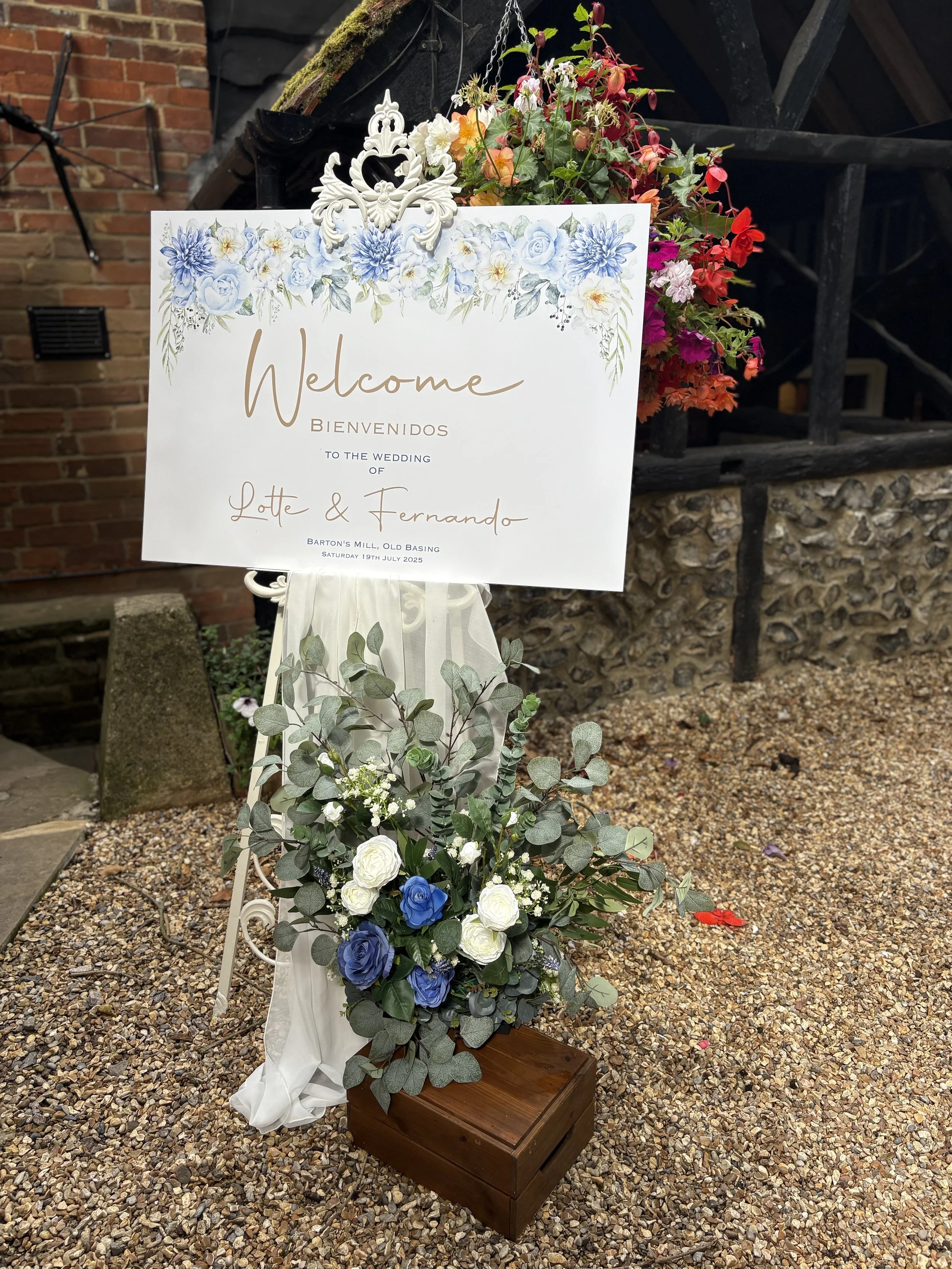 Wedding welcome sign with floral decoration and a flower arrangement on the ground, set outdoors near a brick wall and gravel ground.