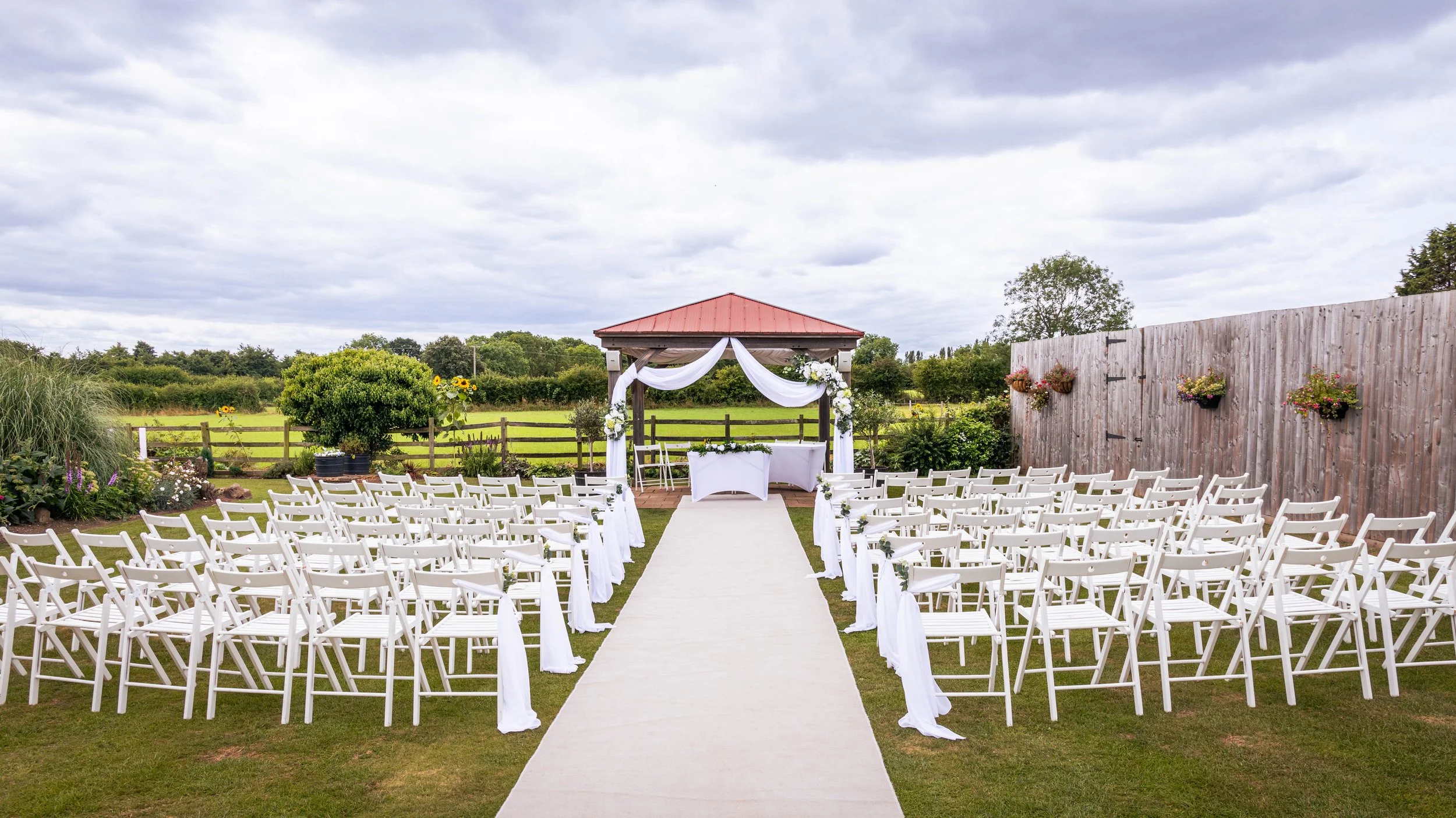 Outdoor wedding ceremony setup with white chairs, a white aisle runner, decorated gazebo, and floral arrangements in a garden with a wooden fence and cloudy sky.
