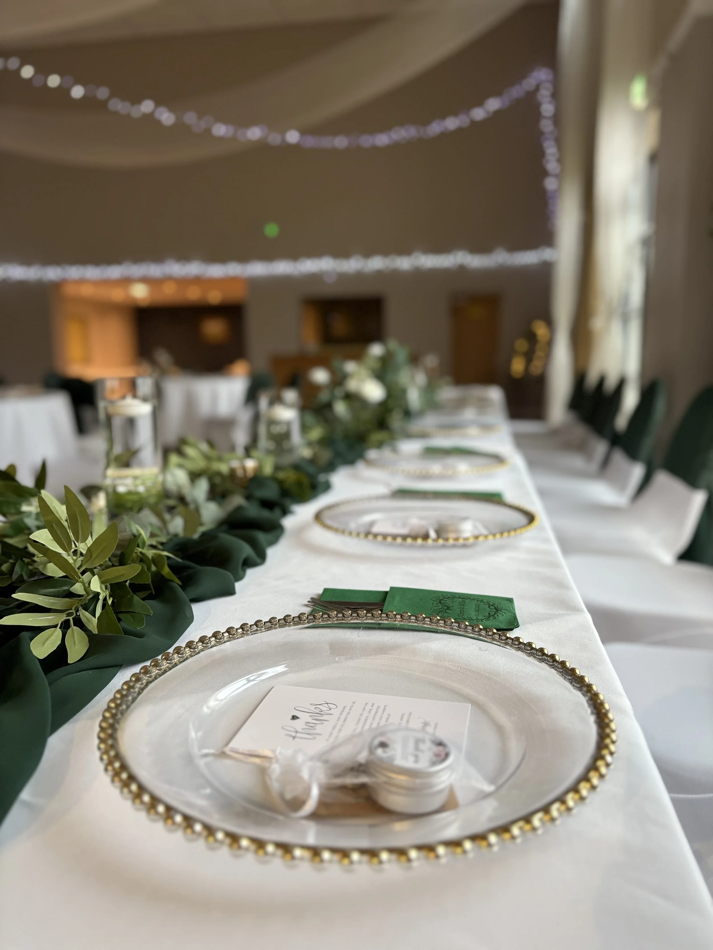 Wedding reception table decorated with greenery, gold-trimmed clear plates, and place cards in an indoor venue with string lights.