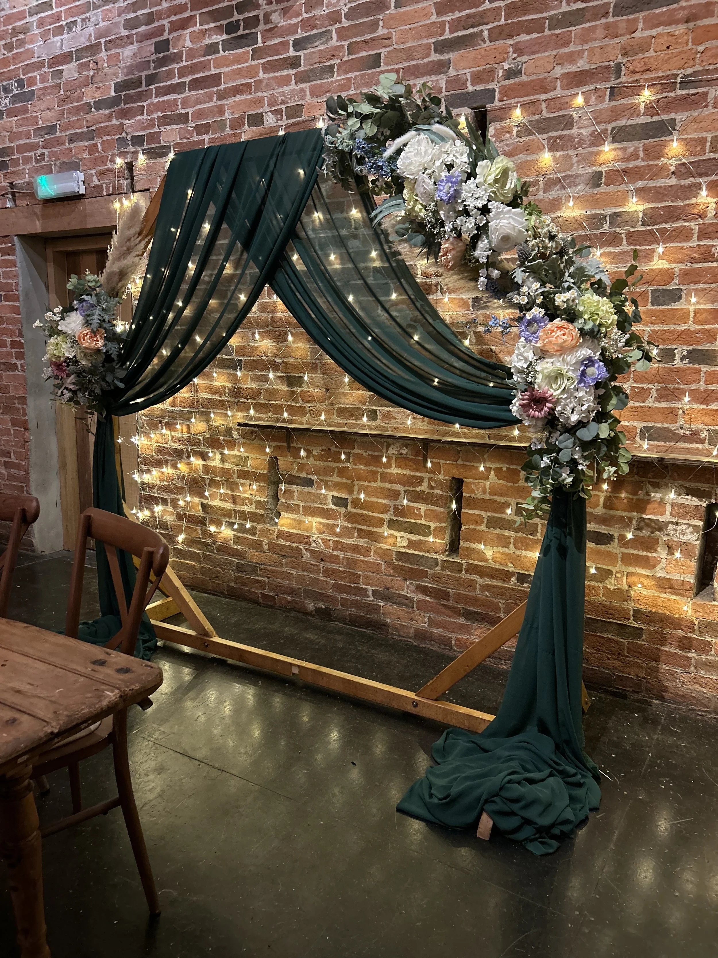 A decorative wedding arch with dark green drapes and a floral arrangement of white, peach, purple, and green flowers, topped with greenery, set against a brick wall decorated with string lights.