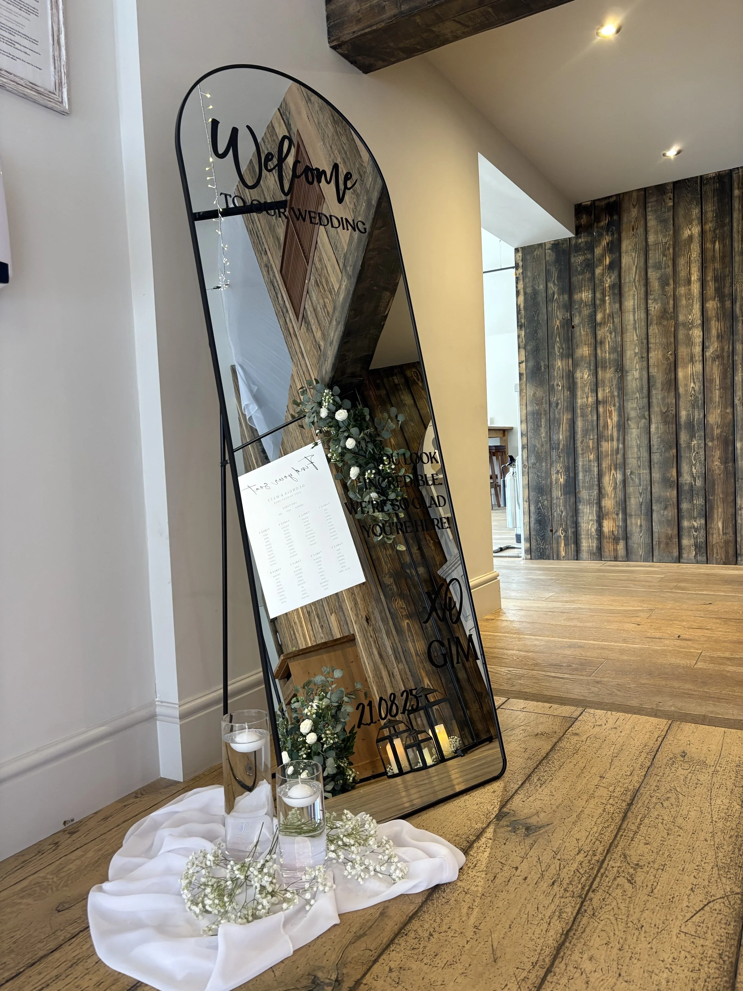 Decorative wedding mirror with writing, floral arrangements, candles, and a table with candles and flowers on a draped cloth, placed on wooden floor near a white wall and a wood-paneled wall.