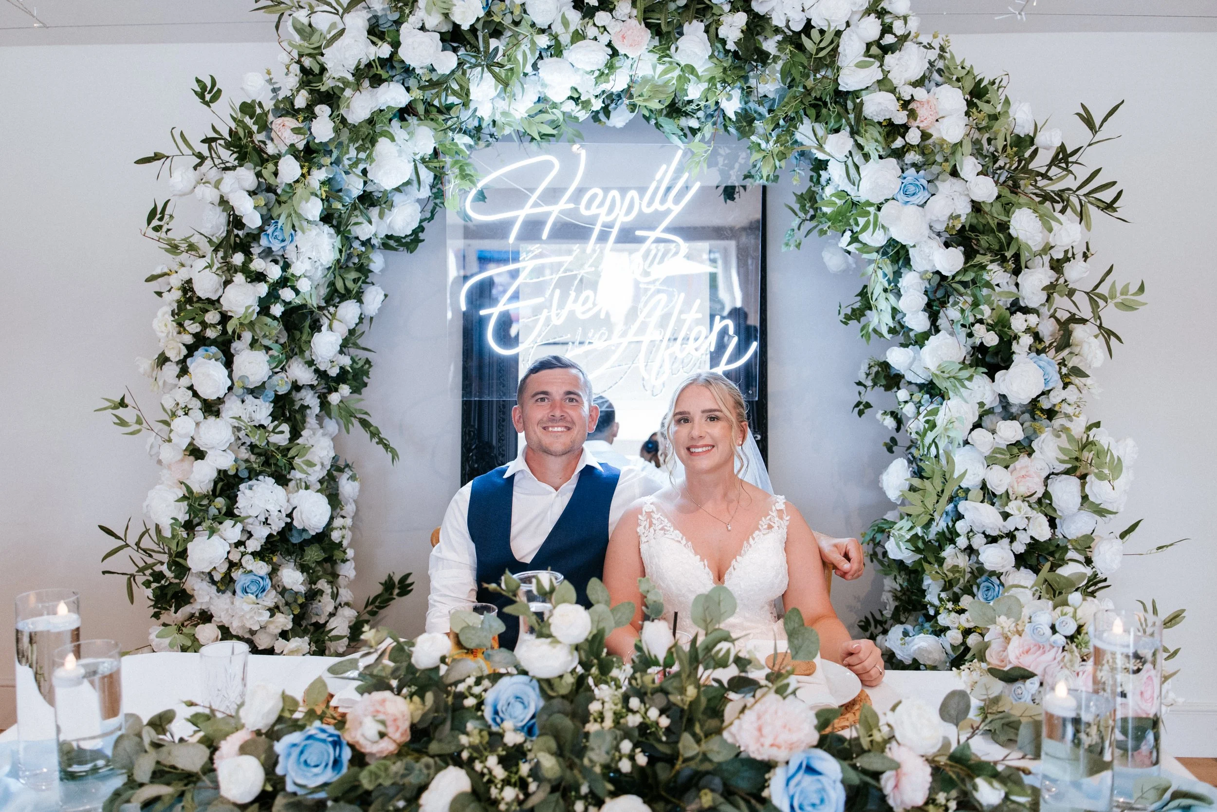 Sweetheart table styled with luxury floral and foliage arch with nuetral colors with pops of blue with a large neon happily ever after sign suspended from the arch with a matching table floral arrangement completed by floating candles