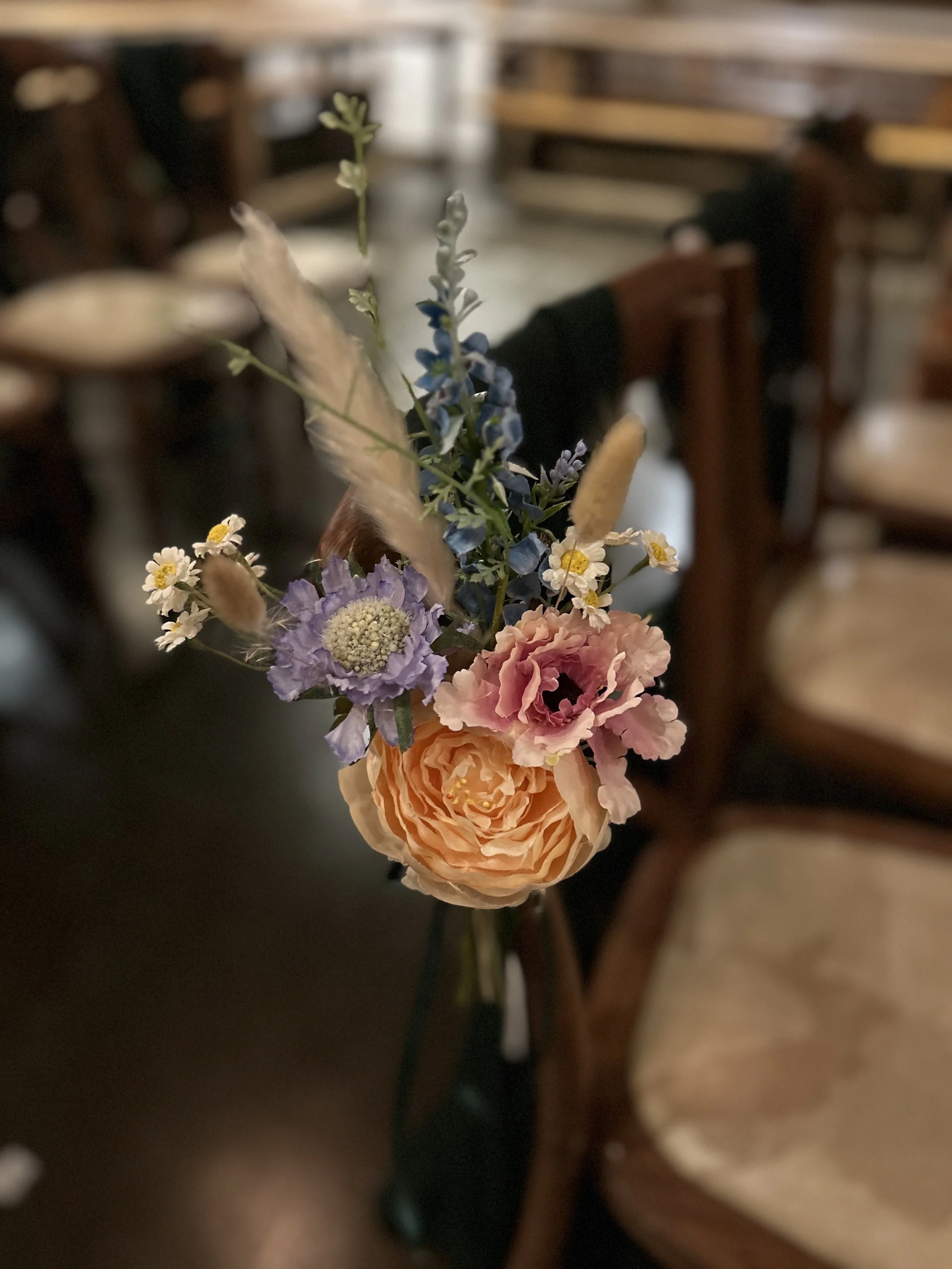 A small bouquet of various flowers including a large peach-colored rose, pink and purple blossoms, small white daisies, and blue flowers, arranged in a narrow black vase.