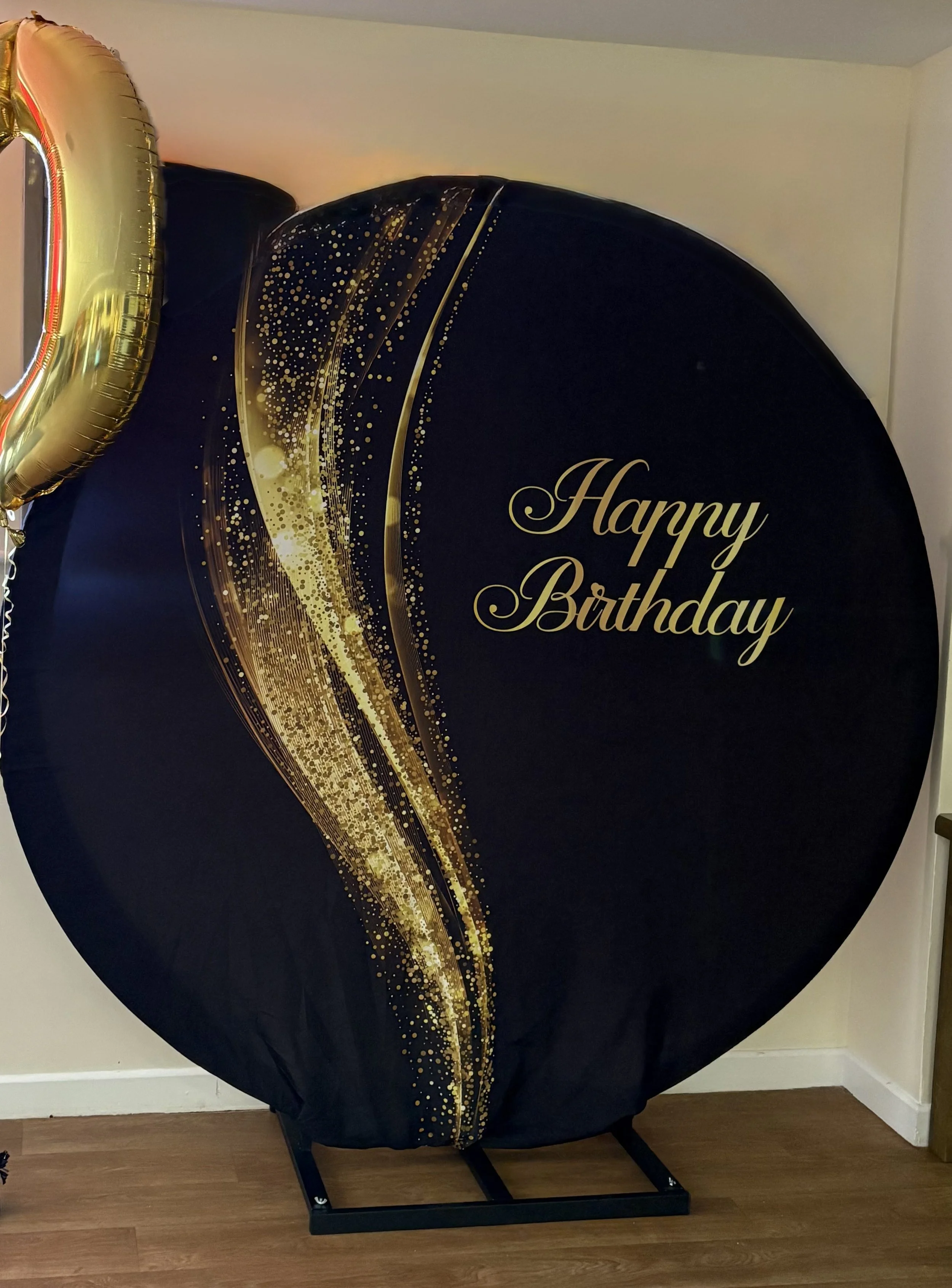 Large black birthday balloon with gold sparkling design and gold cursive text 'Happy Birthday' on it, placed on the floor against a white wall.