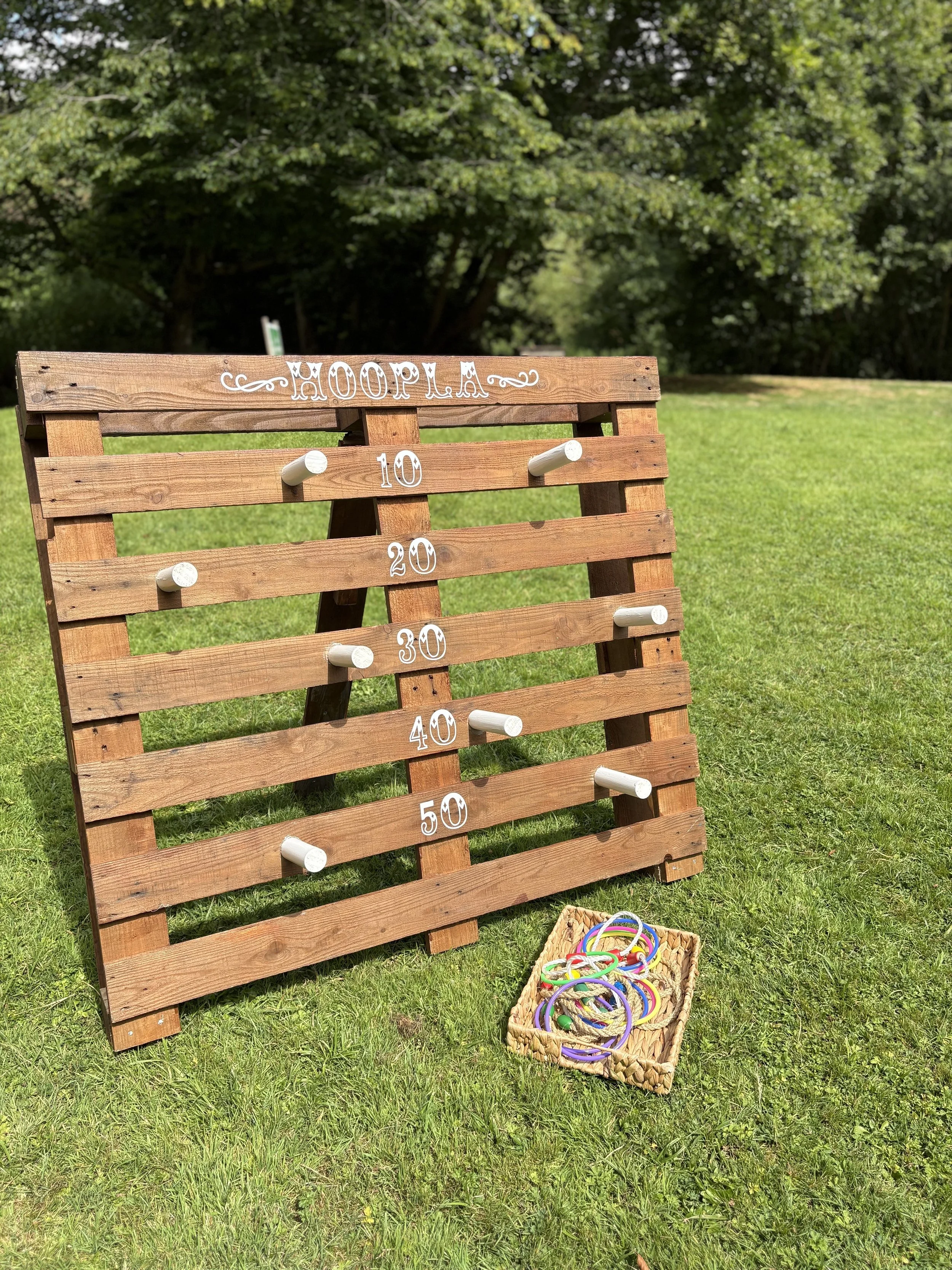 A wooden hoopla game with white pegs and numbered rings on a grassy field, with a basket of colorful rubber bands nearby and trees in the background.