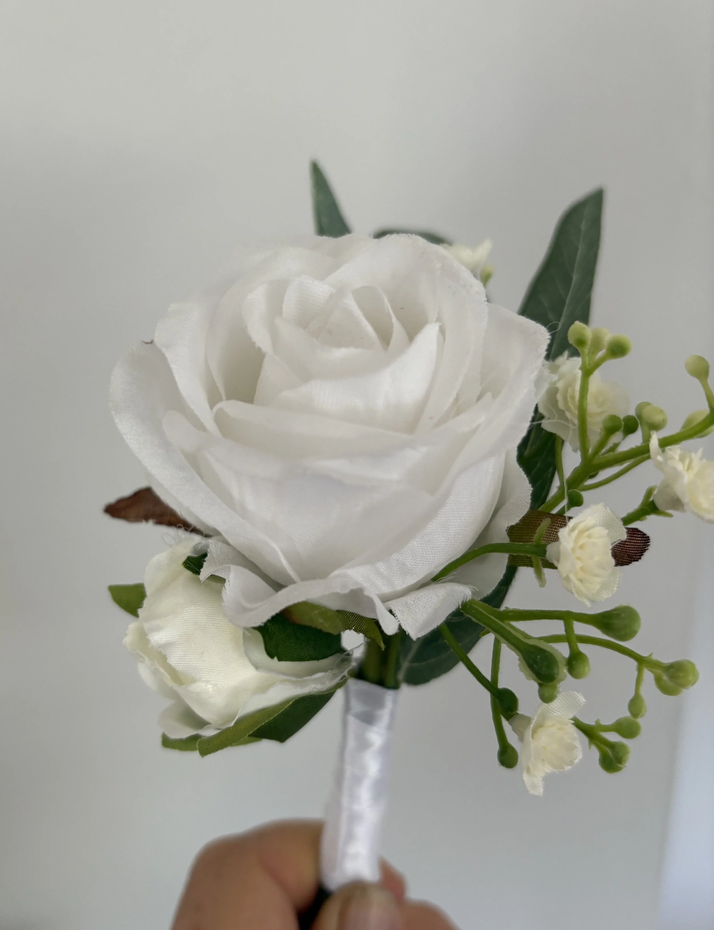 White artificial rose boutonniere with small white flowers, green leaves, and a satin ribbon handle.