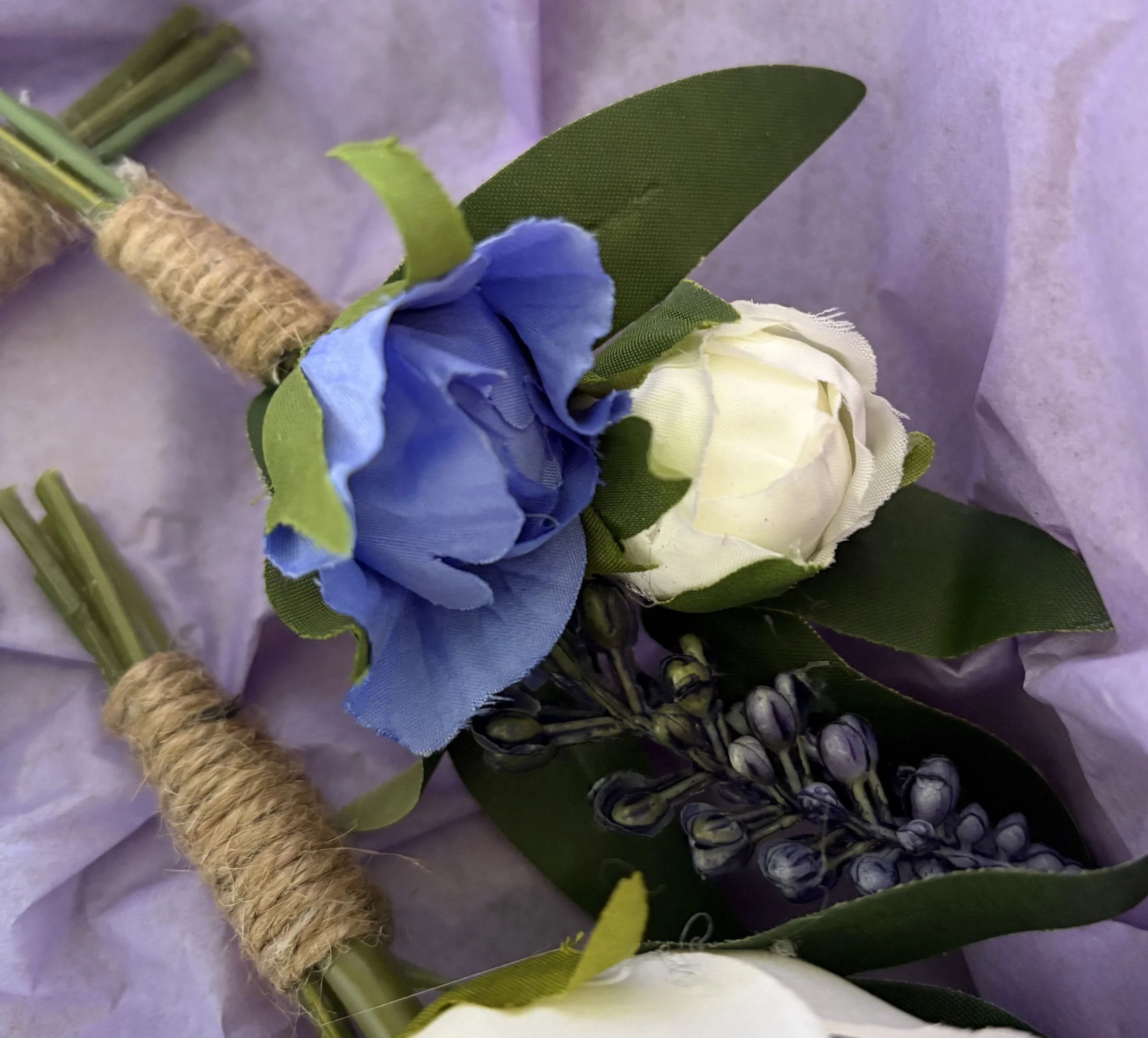 Artificial blue and white boutonniere with green leaves and lavender buds, wrapped with twine, on purple tissue paper.