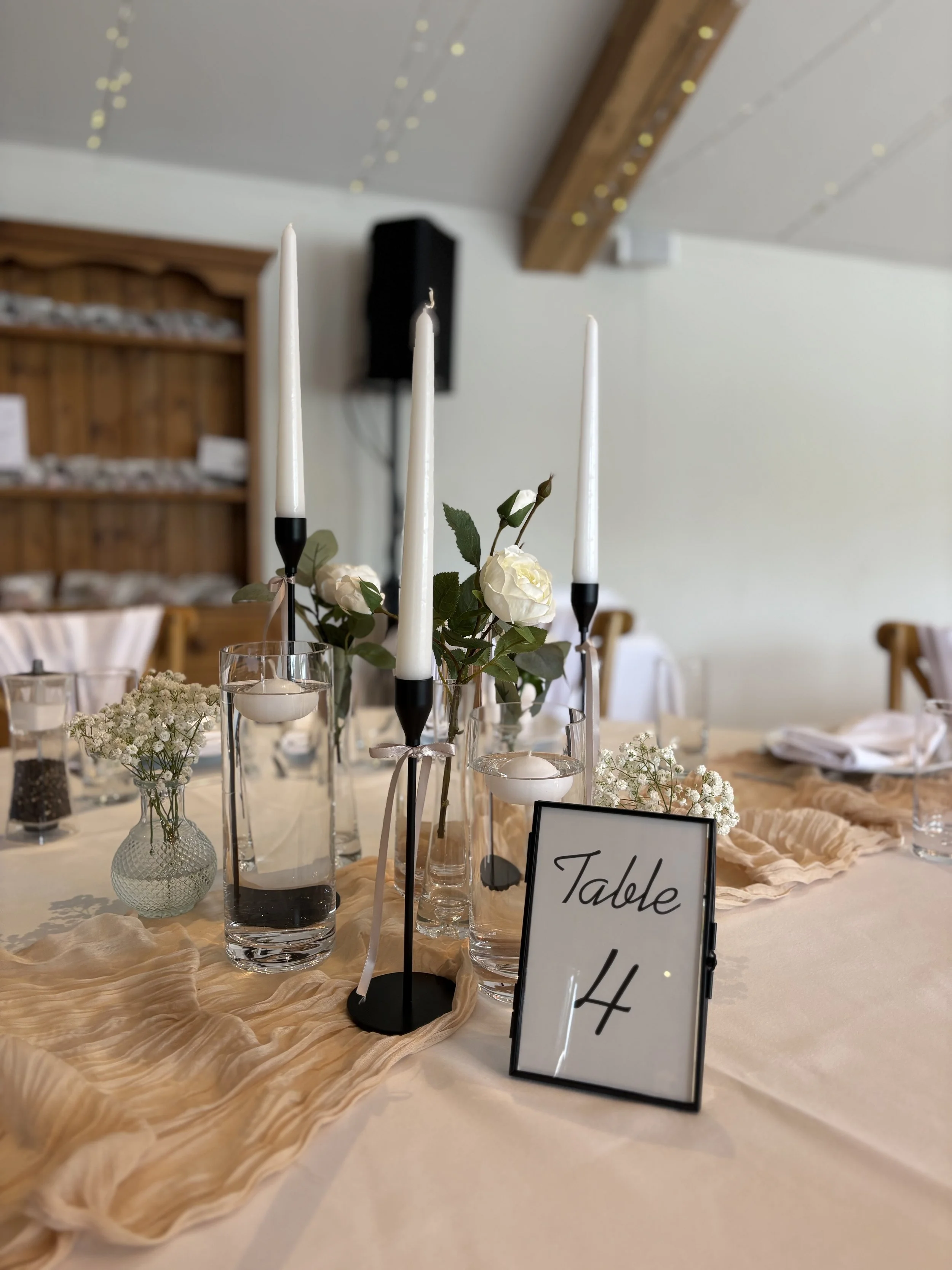 Wedding table decor with white candles, white flowers, glass vases, and a sign labeled 'Table 4' in a decorated reception hall.