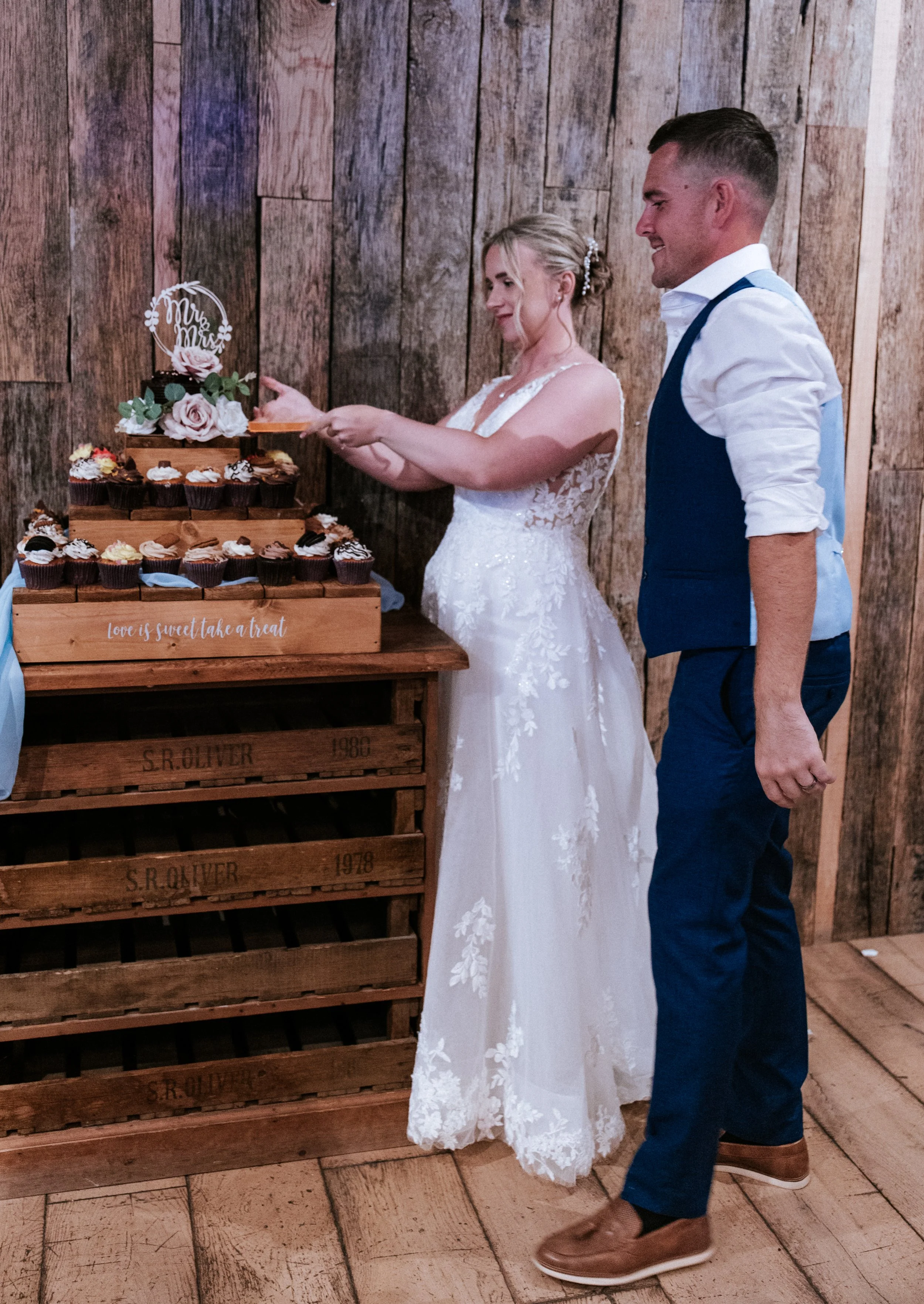 A bride and groom standing by a table with cupcakes, the bride cutting a cupcake and a cupcake topper that says 'Mr & Mrs'.