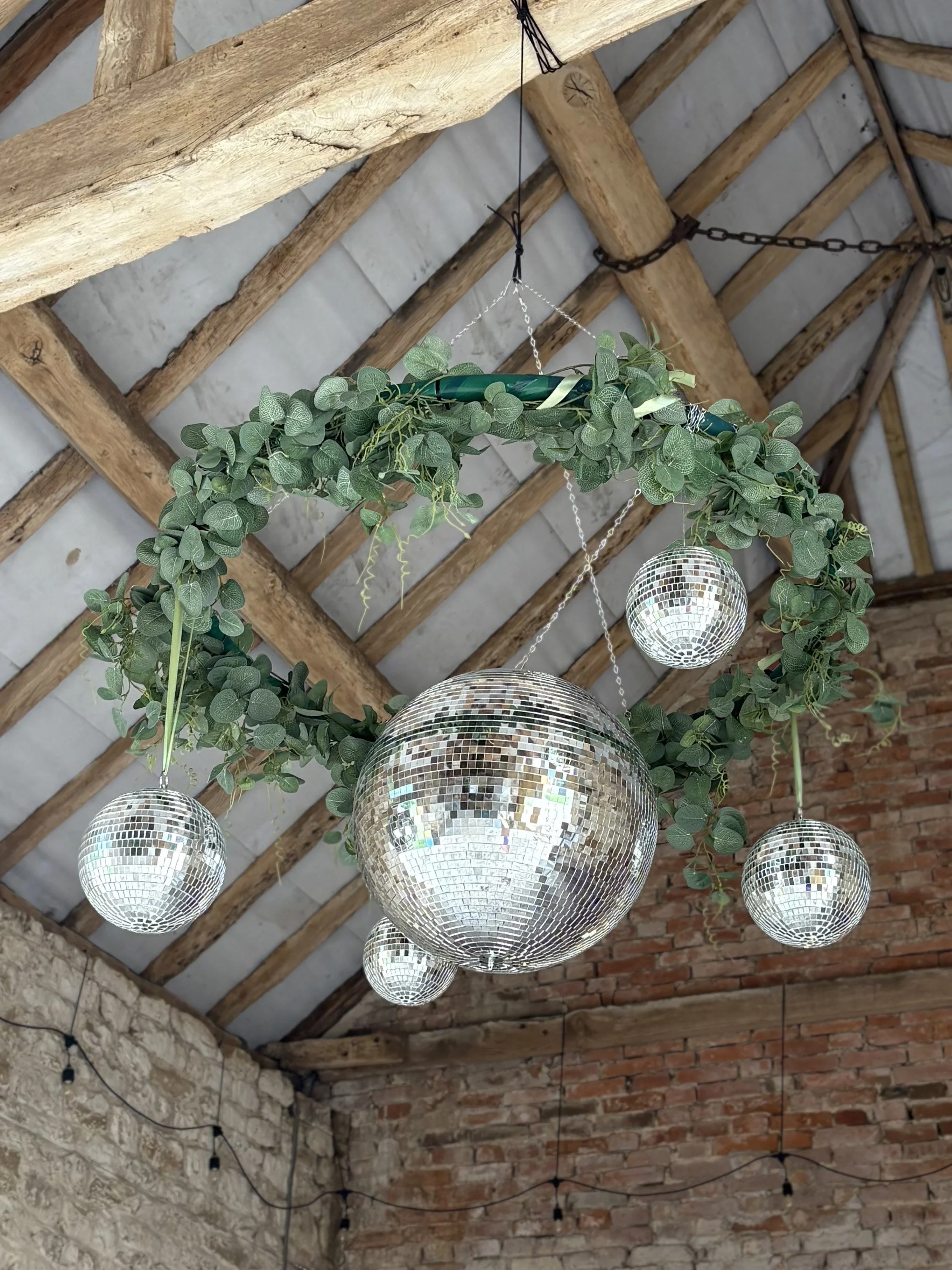 Disco balls hanging from a leafy garland in a rustic, brick and wood ceiling interior.