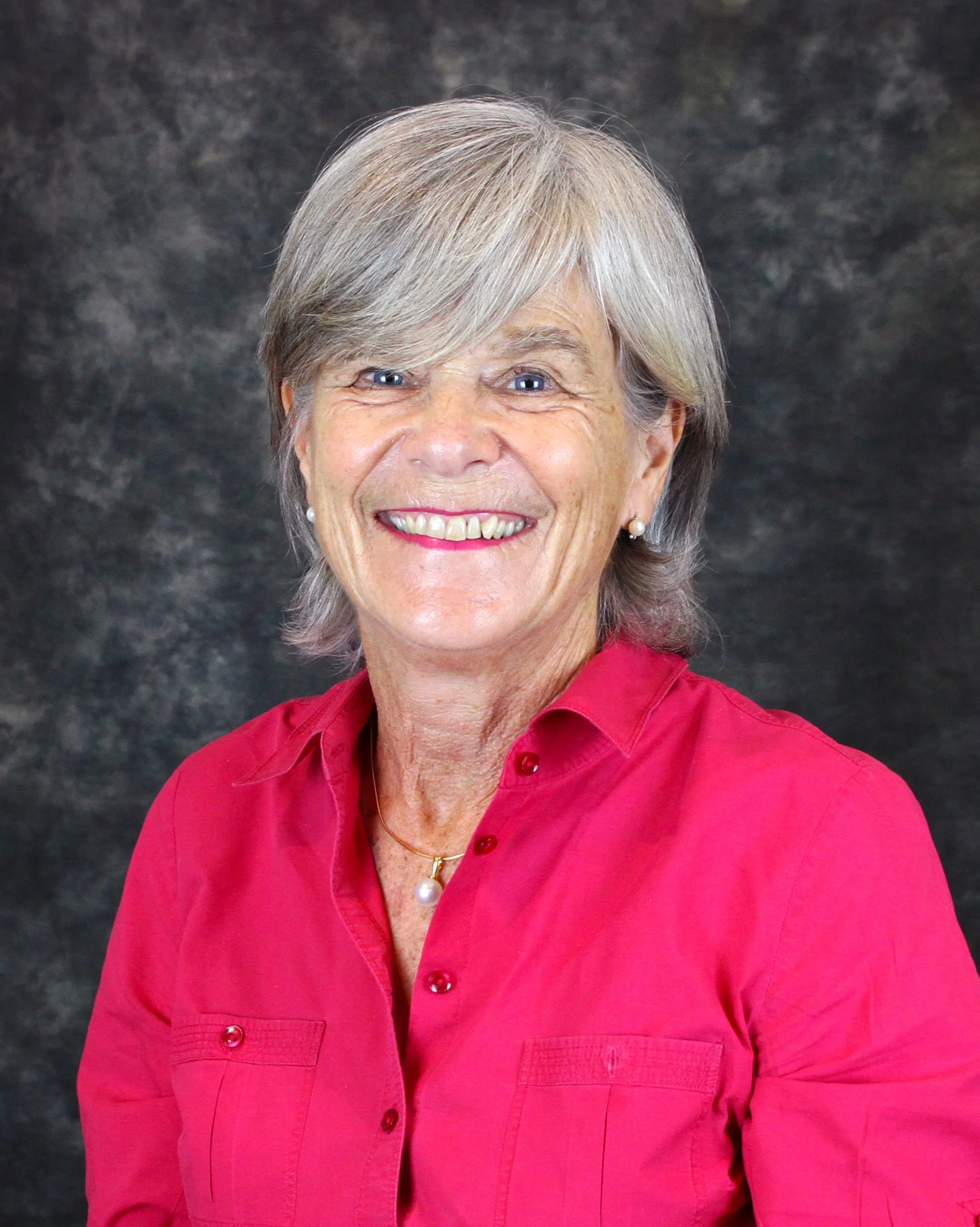 A smiling woman with short grey hair, wearing a red button-up shirt, pearl earrings, and a pearl necklace, against a dark textured background. A trusted friendly marriage celebrant.