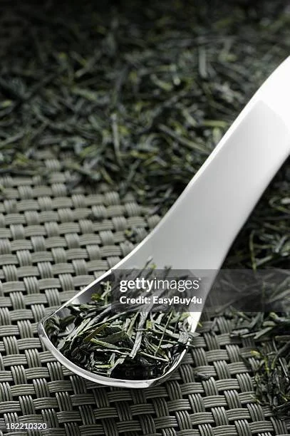 Close-up of a white plastic spoon filled with dried tea leaves, resting on a textured black surface outdoors.