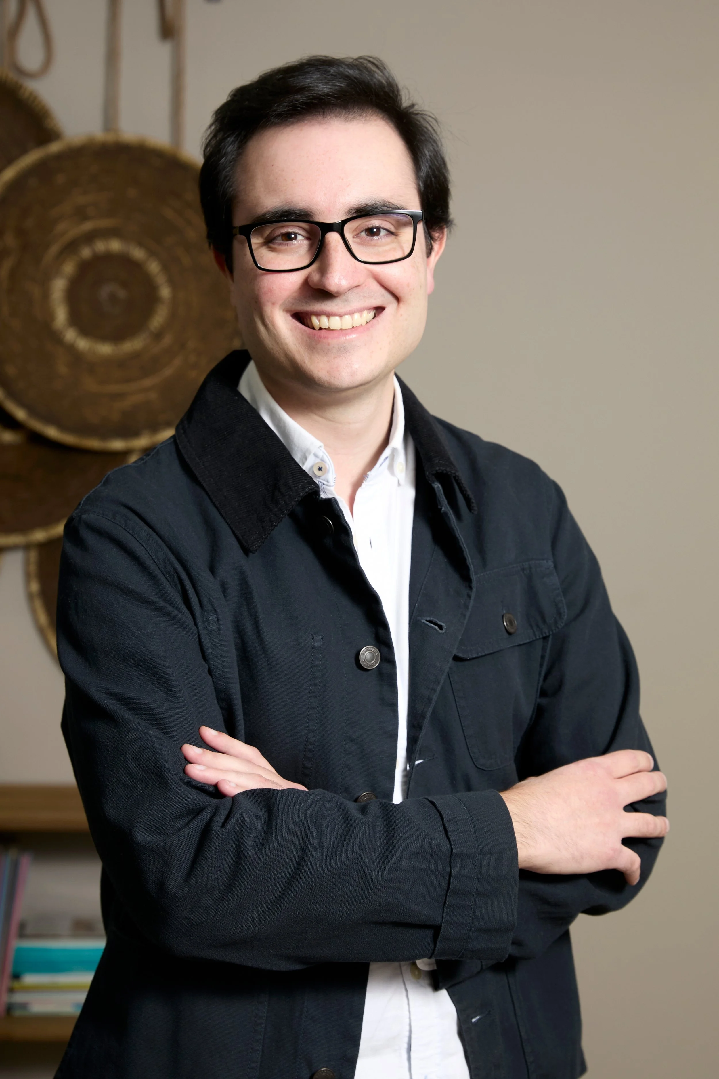 Joven hombre con gafas y ropa oscura, sonriendo, de pie con brazos cruzados en interior con decoraciones de cestas de mimbre en la pared.