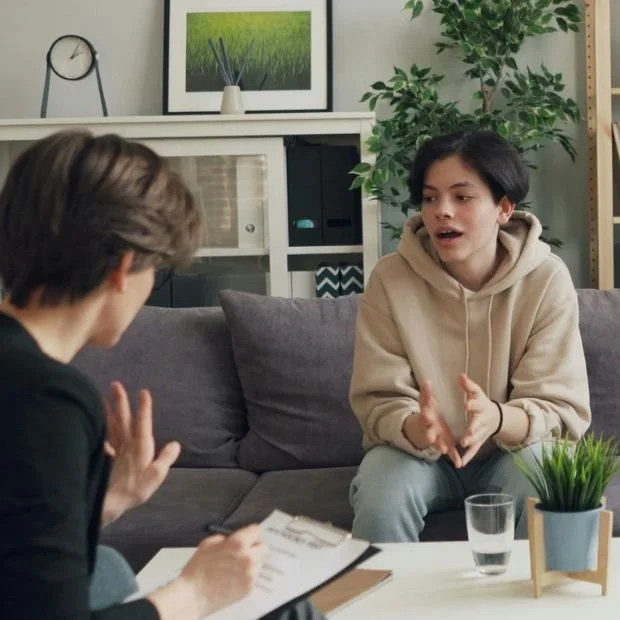 Dos jóvenes conversando en una sala de estar, una mujer con sudadera beige y el cabello corto, y otra persona de espaldas con una libreta, en un entorno acogedor con plantas y decoración moderna.
