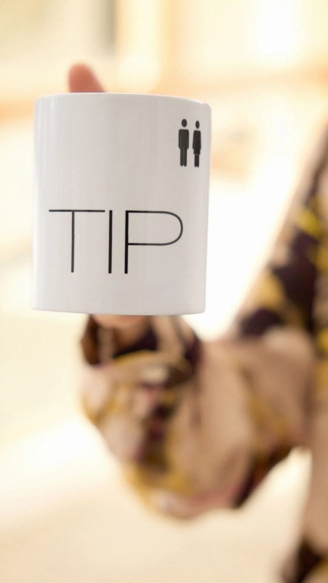 Taza blanca con la palabra 'TIP' y símbolos de hombre y mujer en la parte superior derecha, siendo sostenida por una persona con ropa color beige y detalles en negro y amarillo, en un fondo desenfocado.