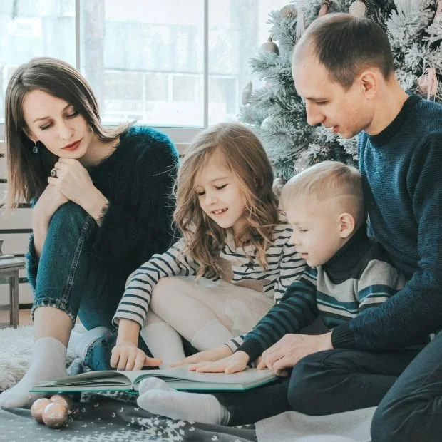 Una familia lee un libro juntos en una sala con un árbol de Navidad decorado.