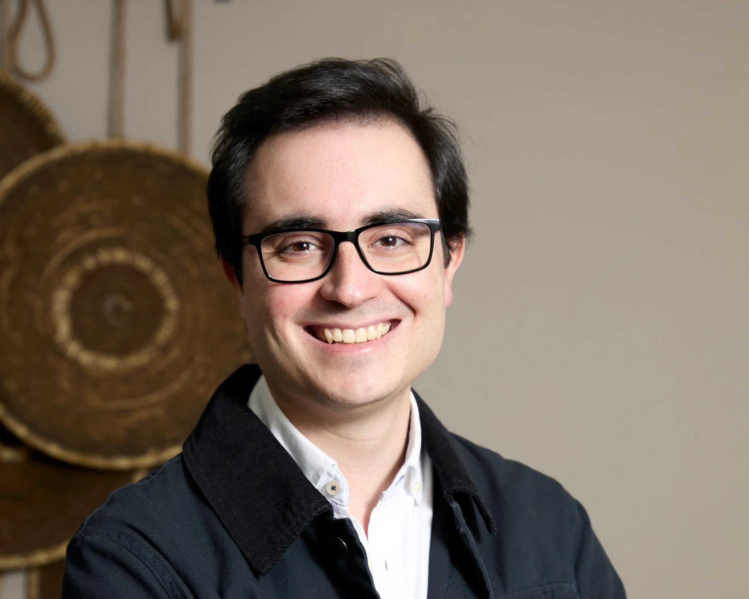 Hombre con gafas y sonrisa, ropa formal, fondo de decoraciones de madera redondas.