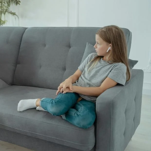 Niña joven con auriculares en el sofá gris, mirando hacia la izquierda, en una sala moderna y luminosa.