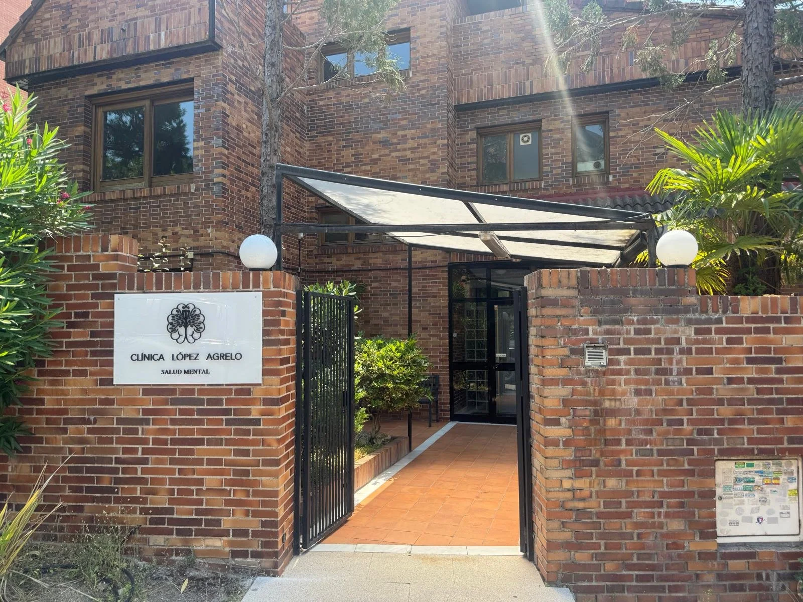Entrada a la clínica López Agoledo, centro de salud mental, con muro de ladrillos y puerta de rejas abiertas, rodeado de plantas y árboles.
