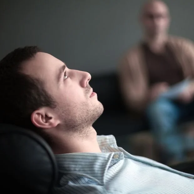 Joven hombre sentado en una sala de terapia, mirando hacia arriba con expresión pensativa, con otra persona al fondo en un sillón.