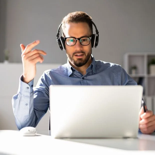 Hombre con gafas y auriculares frente a una computadora portátil en una oficina moderna.