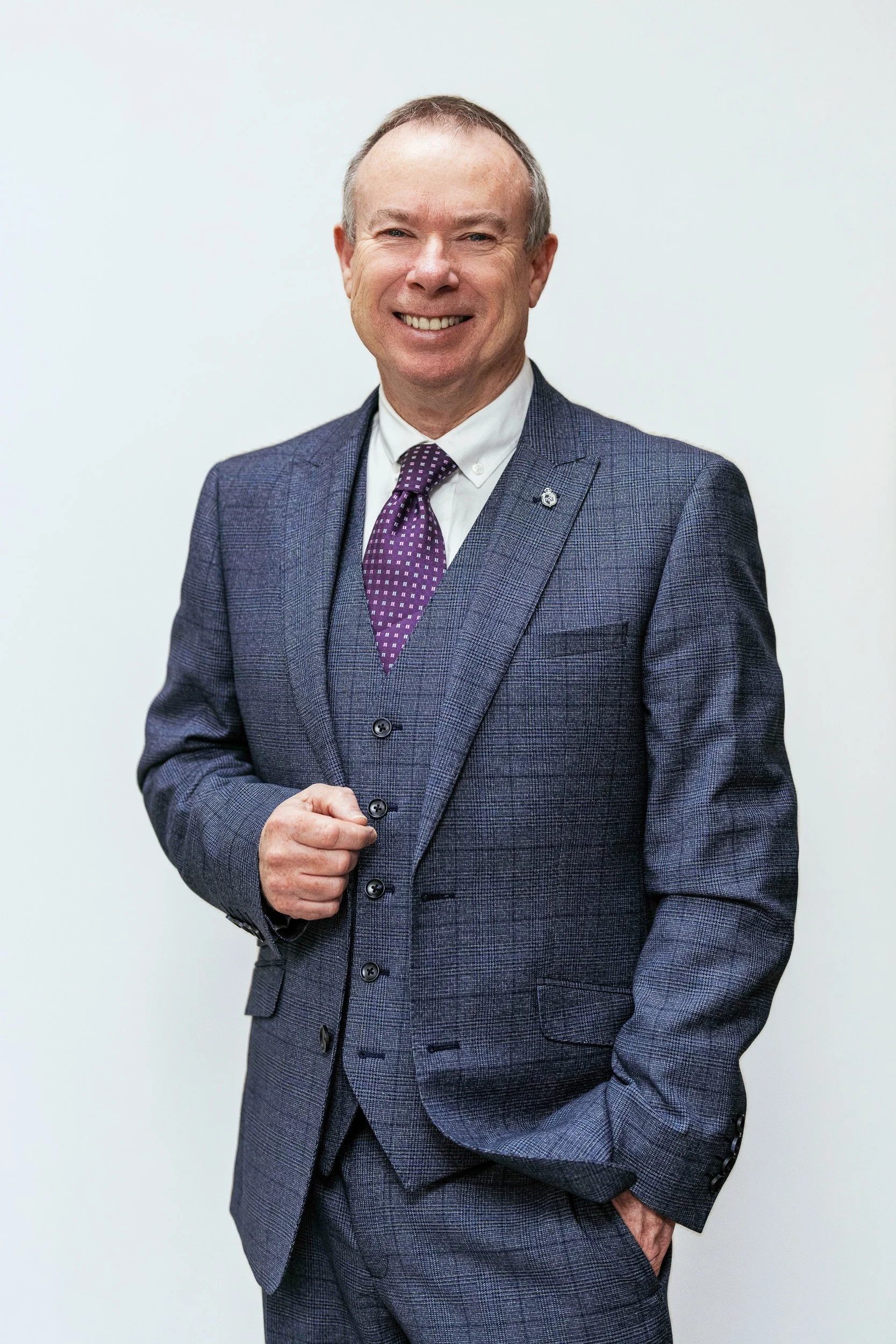Profile photograph of David James Wedding Celebrant dressed in a dark blue plaid suit, white shirt, and purple tie, smiling in front of a plain light-colored wall.