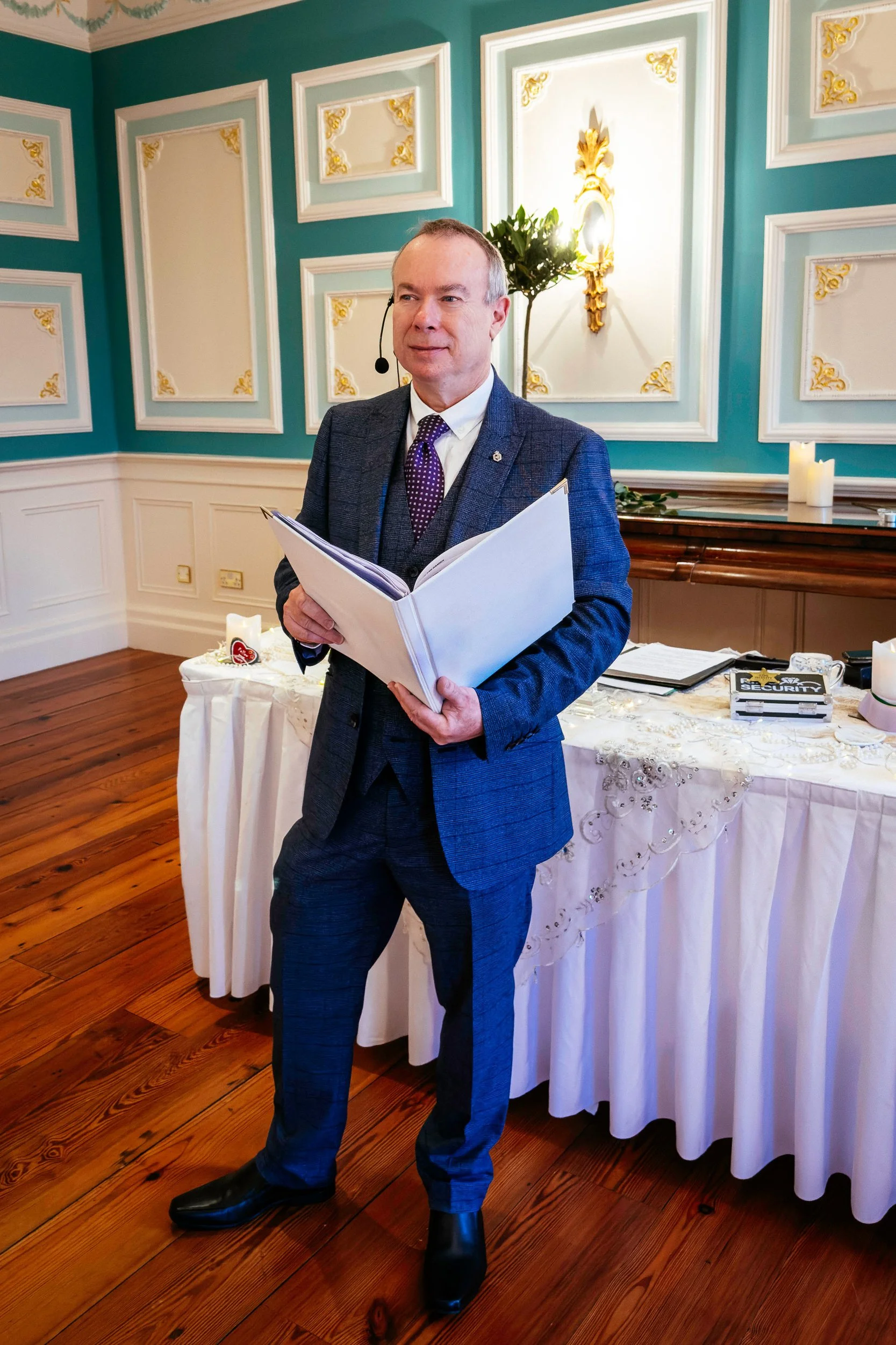 David James  performing a wedding ceremony in the Killashee House Hotel, Naas. He is in a blue suit holding a large open folder stands in a decorated room, with a table behind him covered with a white cloth and candles.
