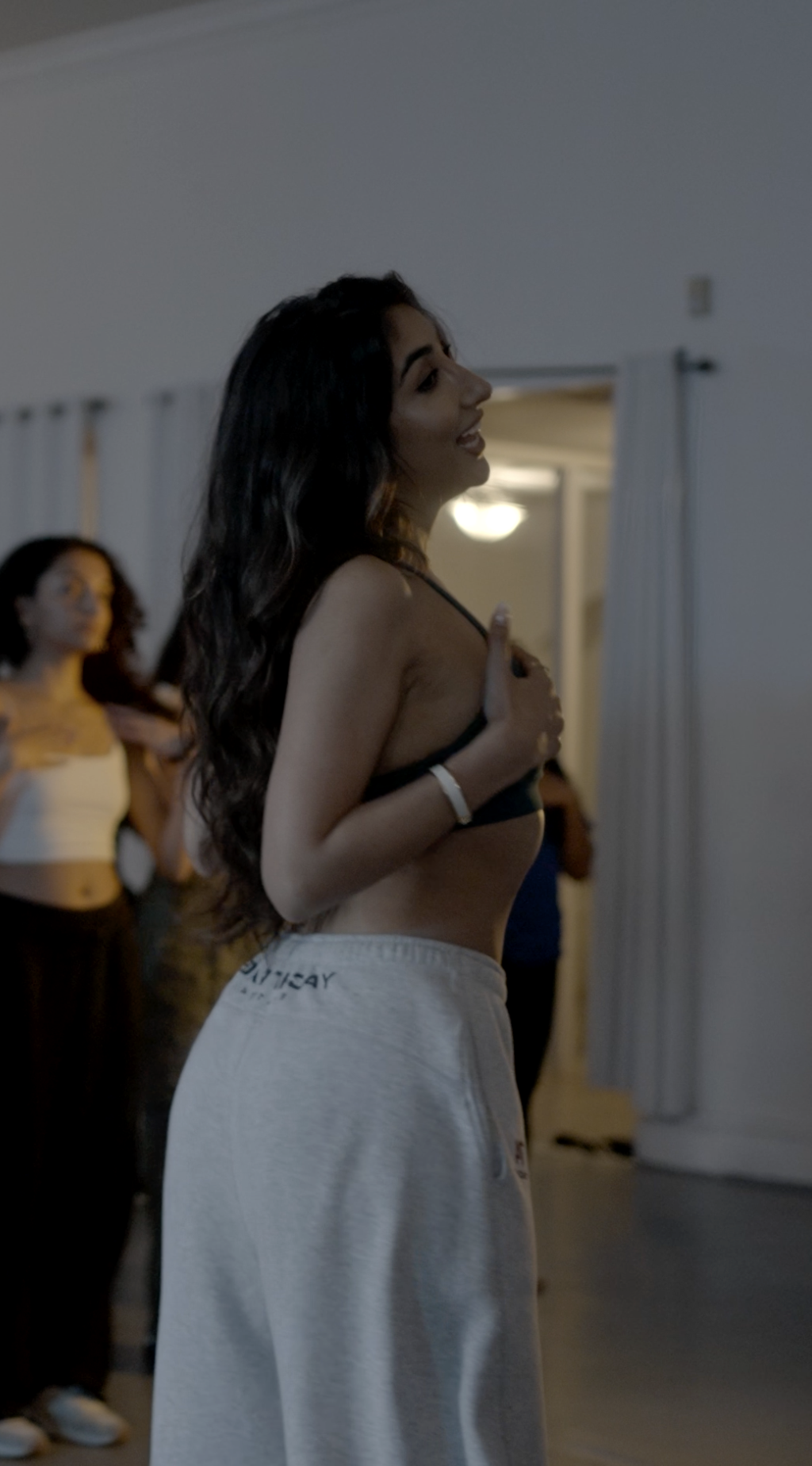 A young woman in a black bra and grey sweatpants stands with her hand on her chest in a dimly lit room, possibly singing or performing. Two other women are in the background, one adjusting her hair.