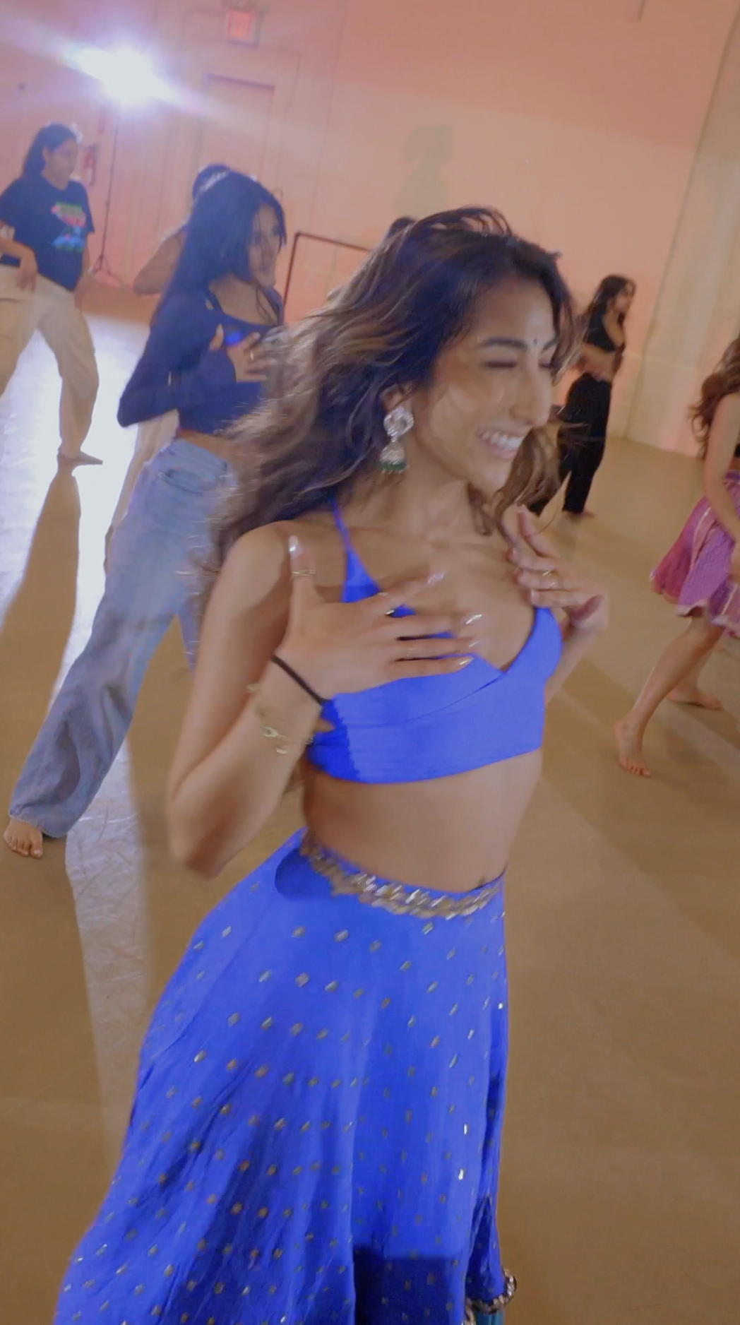 A woman wearing a bright blue crop top and matching long skirt dancing with a joyful smile at a party or celebration indoors with other women in the background.