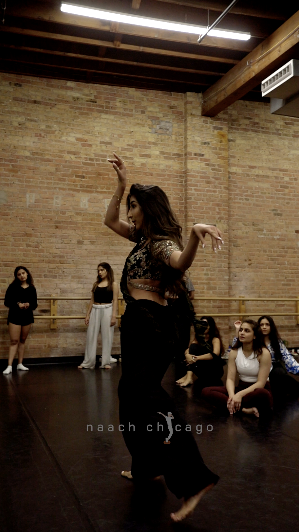 A woman dancing in front of an audience of young women in a dance studio with exposed brick walls and wooden ceiling beams.