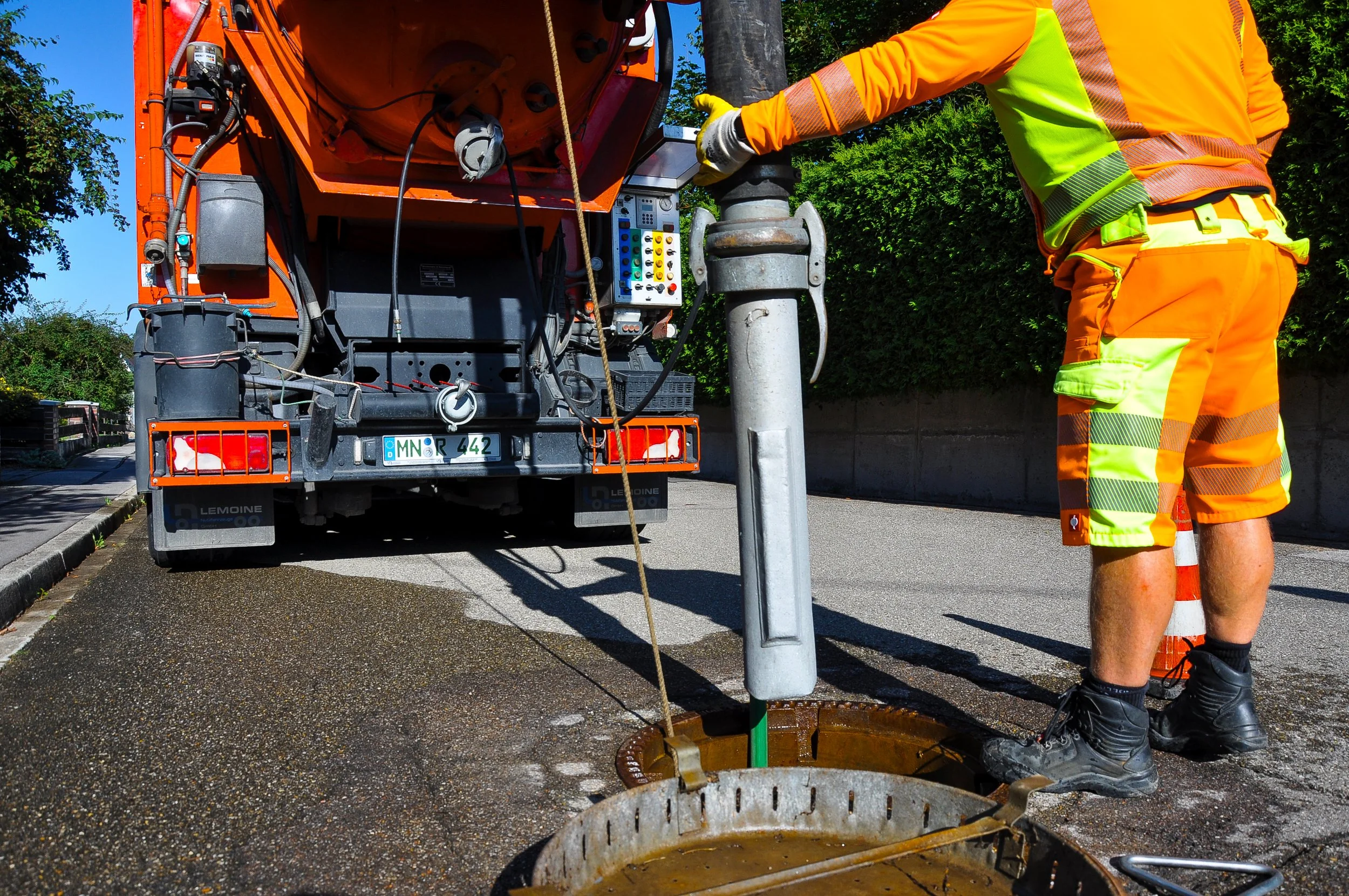 Ein Arbeiter in orange-gelber Warnkleidung arbeitet an einer Straßenlaterne, die in eine  Straßenbohrung eingebaut wird, mit einem orangefarbenen Fahrzeug im Hintergrund.