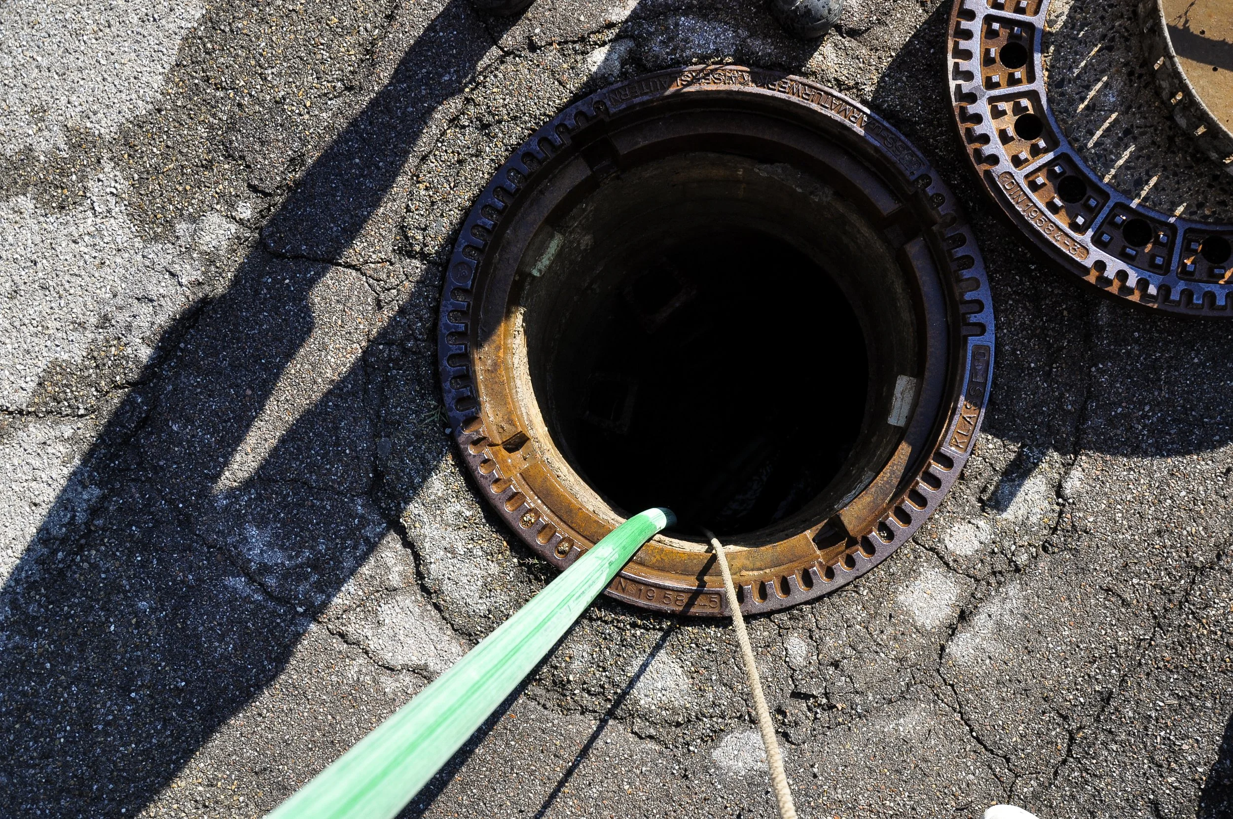 Ein offener Kanaldeckel auf einer asphaltierenden Straße, mit einem grünen Schlauch darin. Es sind zwei Schatten auf dem Boden sichtbar, und ein Seil ist an dem Kanaldeckel befestigt.