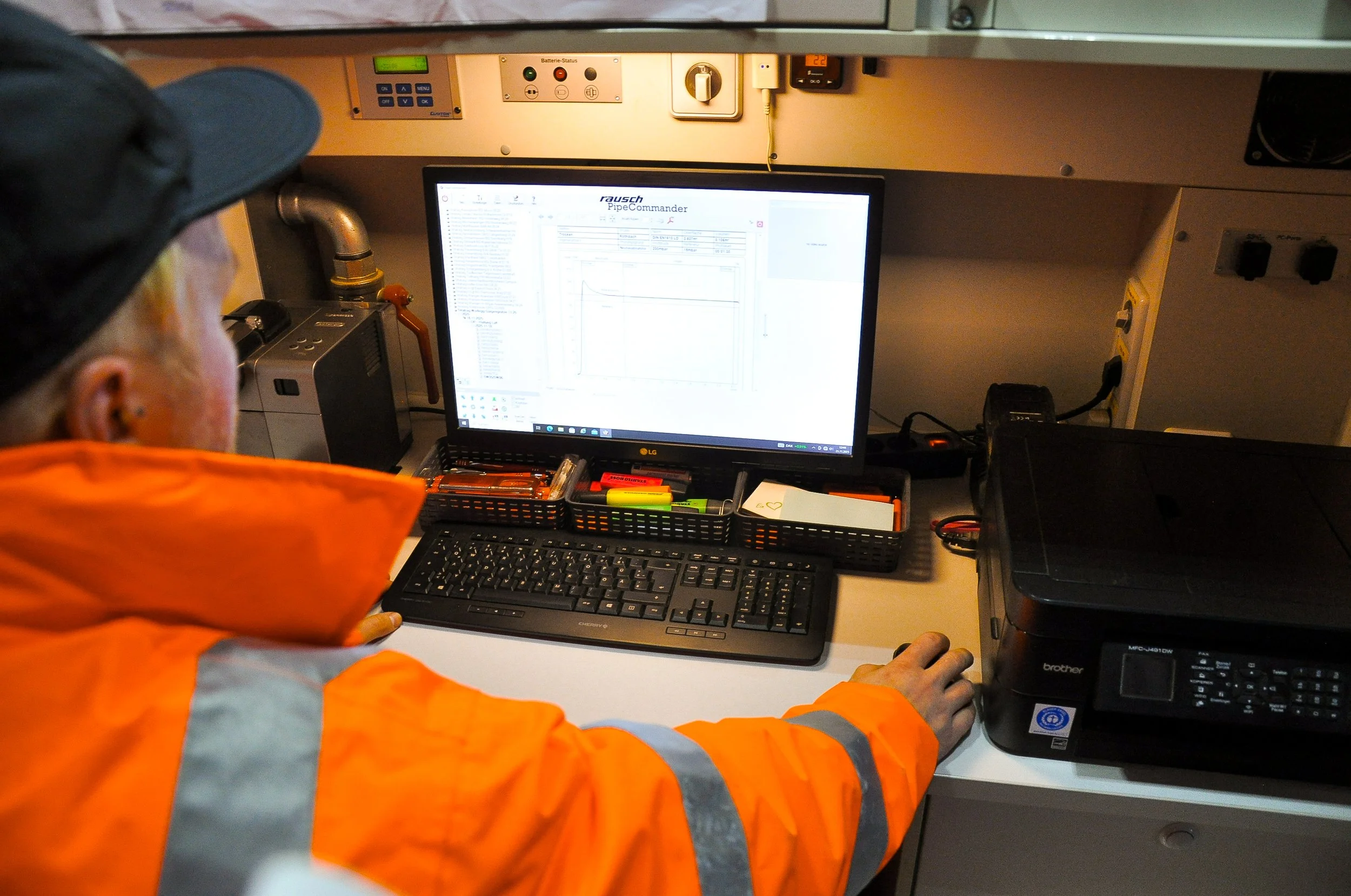 Mitarbeiter in orange Sicherheitskleidung arbeitet an einem Computer mit Diagrammen auf dem Bildschirm, umgeben von Büromaterialien und technischen Geräten.