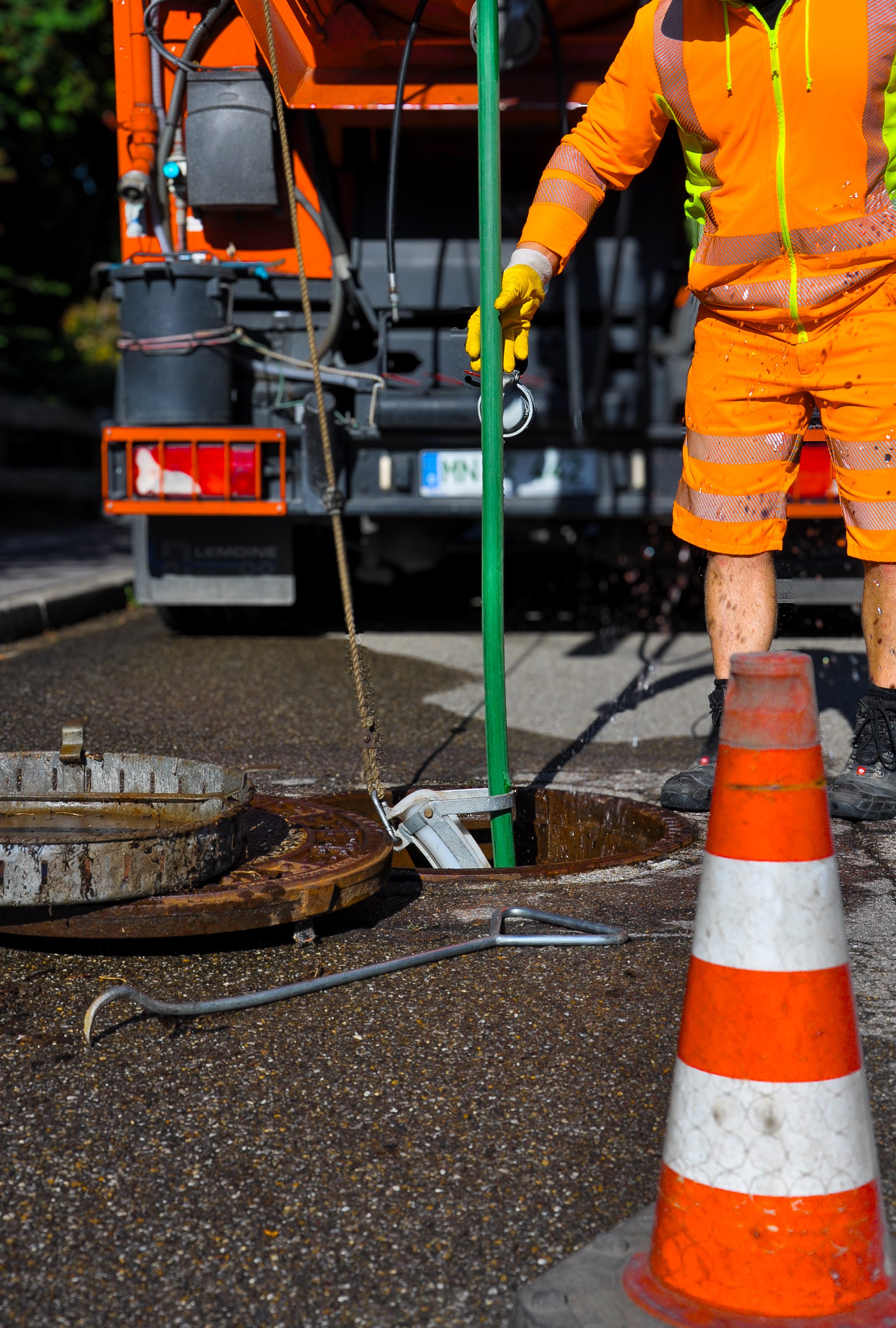 Arbeiter in orange Warnkleidung repariert eine Straßenleitung. Ein orangefarbenes Verkehrs- oder Sicherheitshütchen steht im Vordergrund.